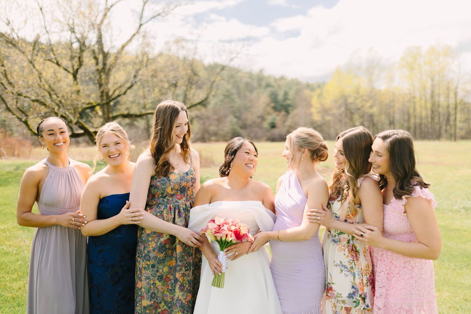 Bride and bridesmaids in mismatched dresses laughing together outdoors with colorful bridal bouquet in spring setting.
