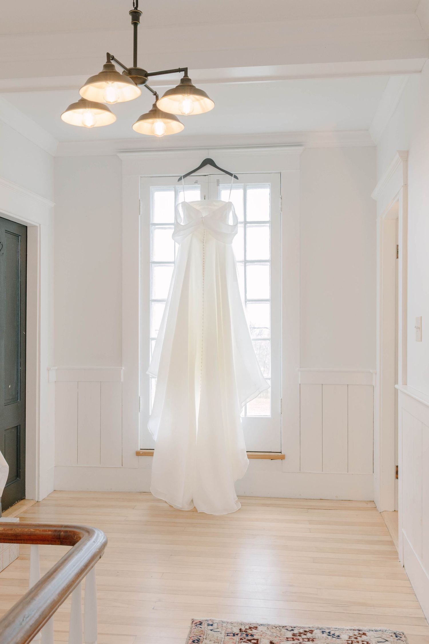 A white wedding dress hanging on a hanger in front of a bright window in an elegant white room with hardwood floors.
