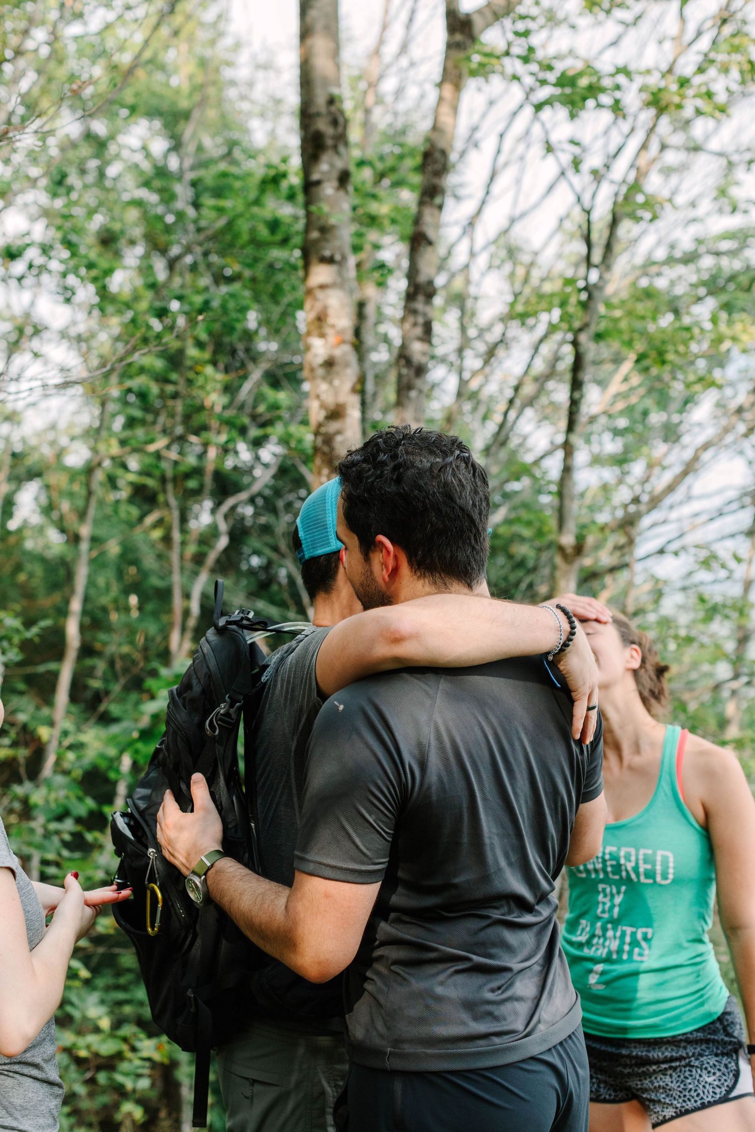 Two people share a warm hug on a forest trail, with a woman in a green tank top nearby.
