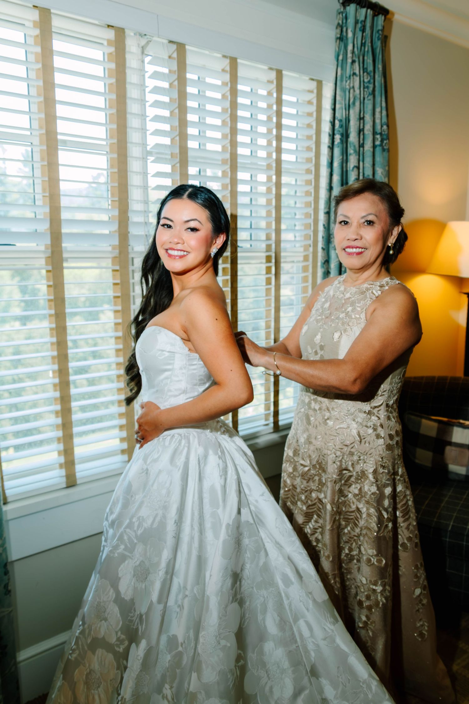 Smiling bride and her mother posing together as mother fastens the back of the wedding gown.