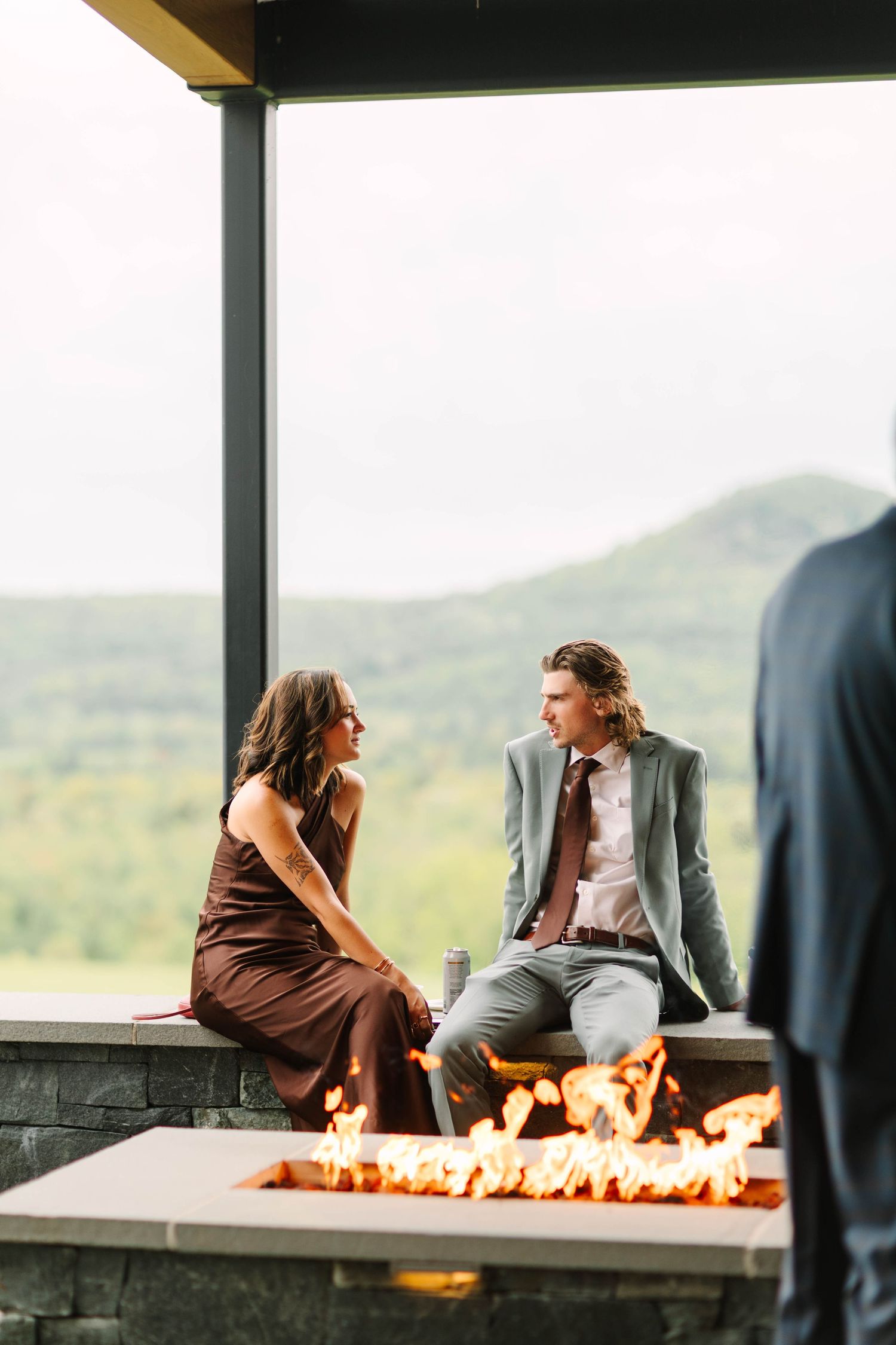 Couple in formal attire sits and chats by an outdoor fire pit with scenic mountain views in the background.