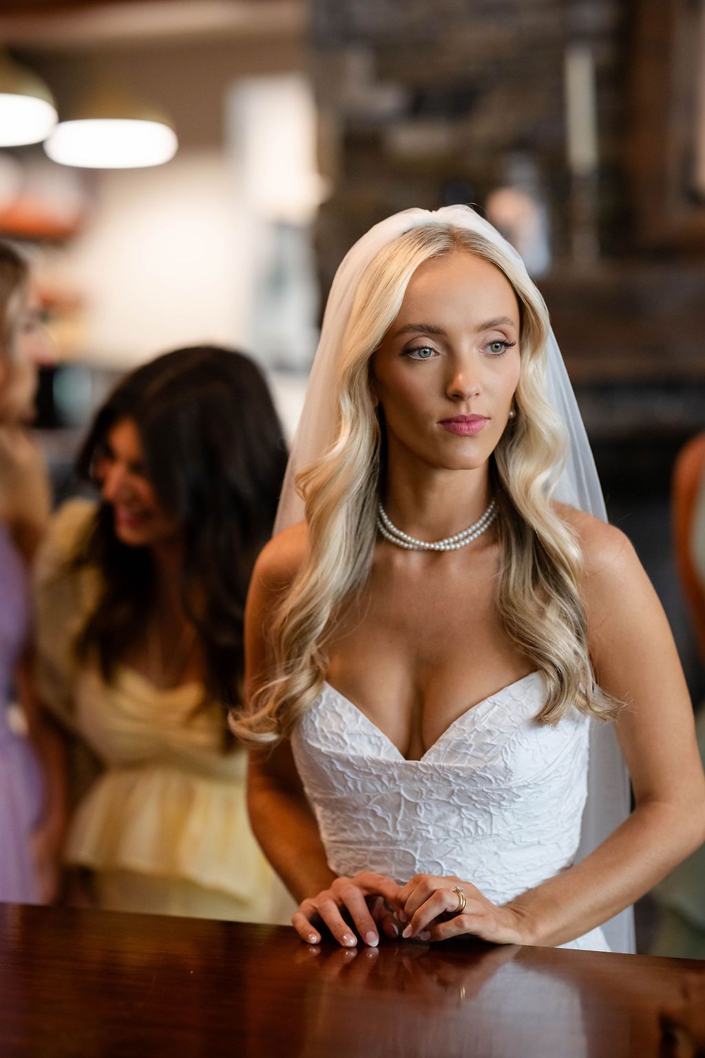 Beautiful blonde bride in a strapless lace wedding dress and pearl necklace leaning against a wooden bar.