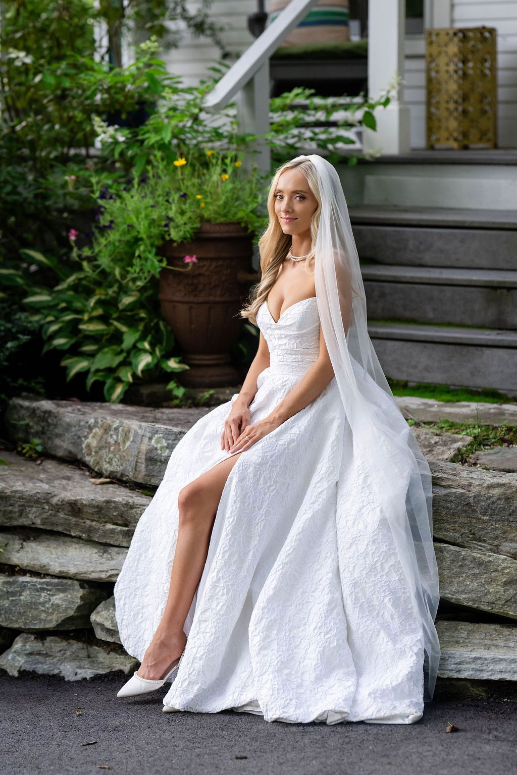 Elegant bride in white lace gown and veil sitting on stone steps surrounded by lush garden greenery.