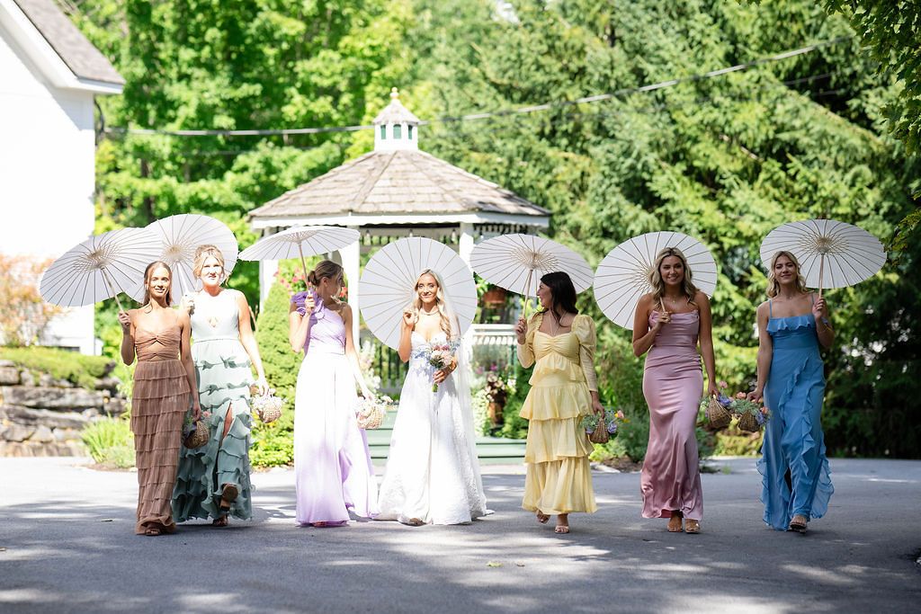 Bride and bridesmaids in colorful mismatched gowns walk with white parasols near a garden gazebo.