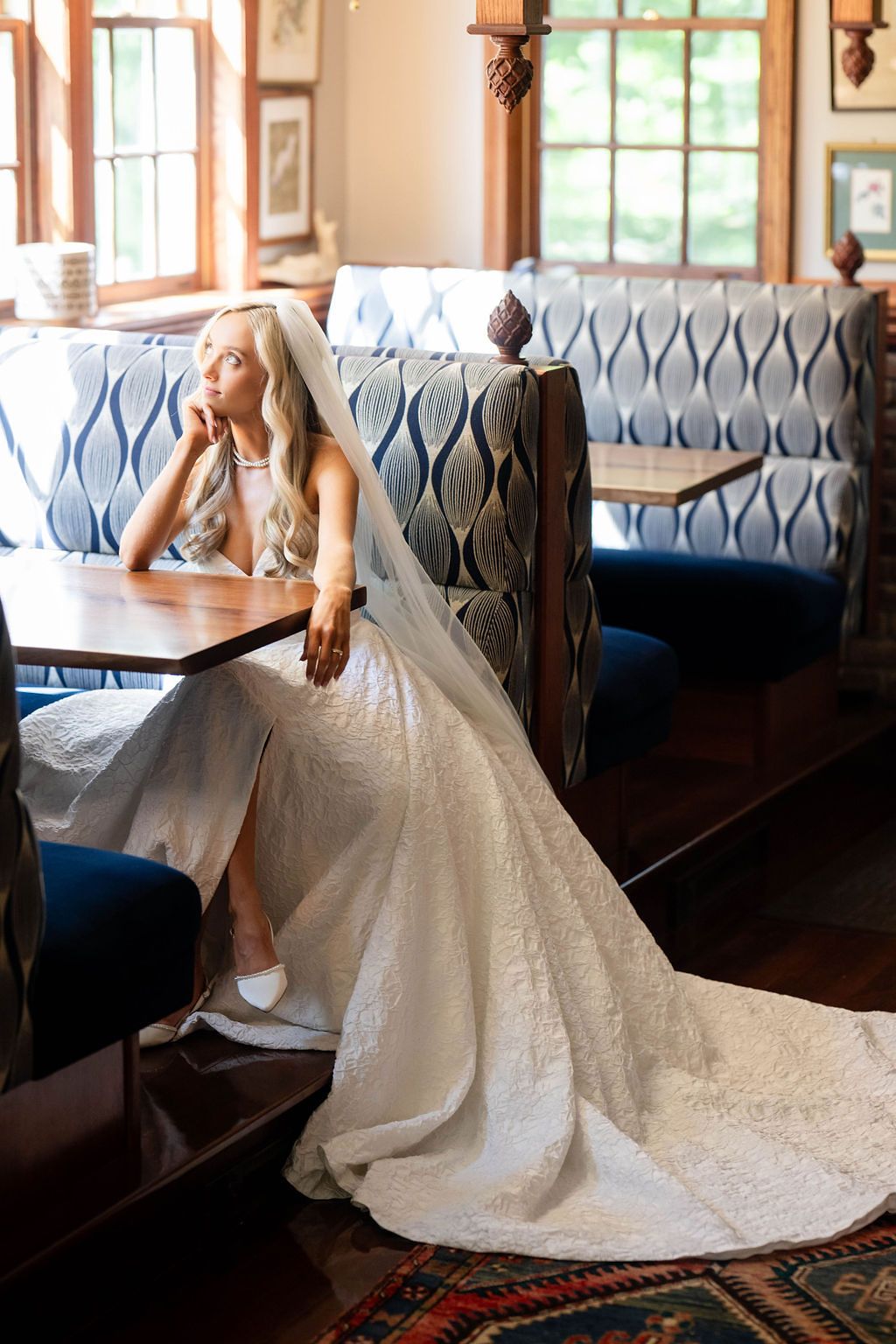 Bride in a lace wedding dress and veil gazes out a window while seated in a blue upholstered booth.