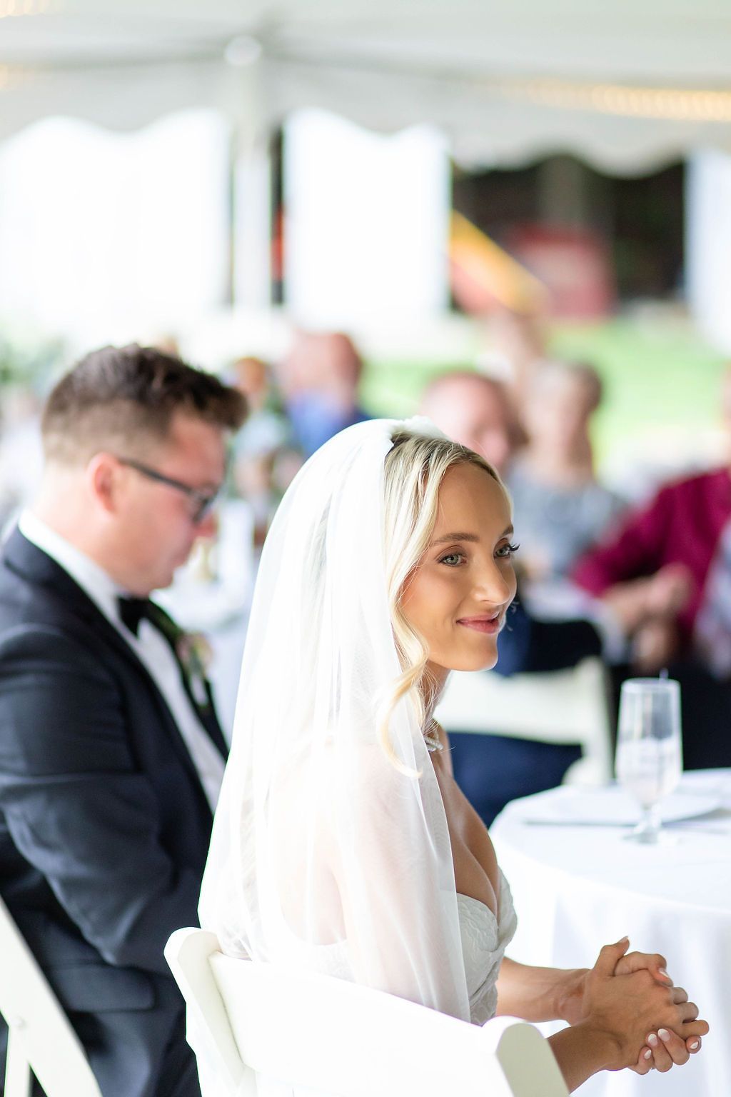 Beautiful blonde bride in white veil smiles softly at reception table during wedding celebration.