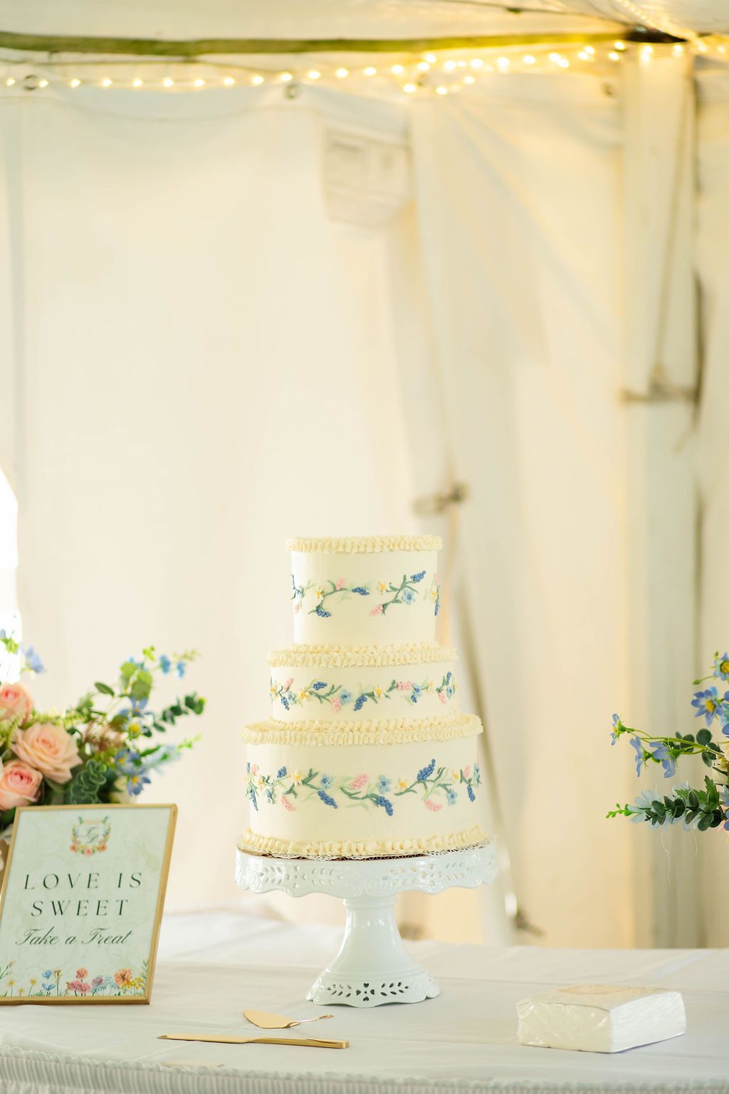 Elegant four-tier white wedding cake with floral details displayed on a cake stand beside a love is sweet sign.