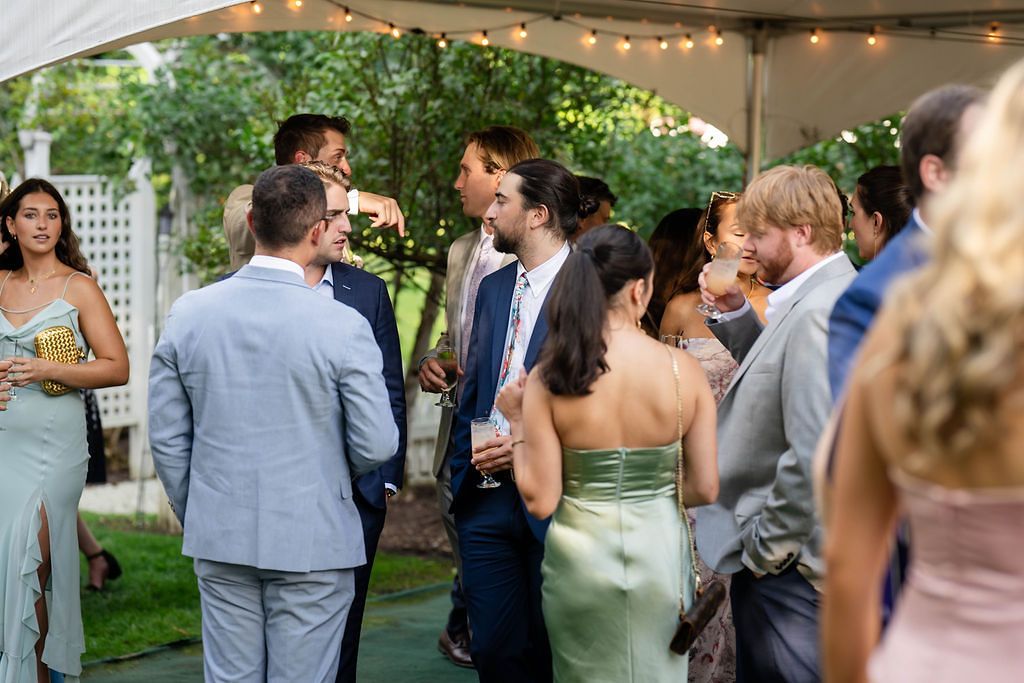 Wedding guests mingling and socializing under a white tent with string lights at an outdoor reception.