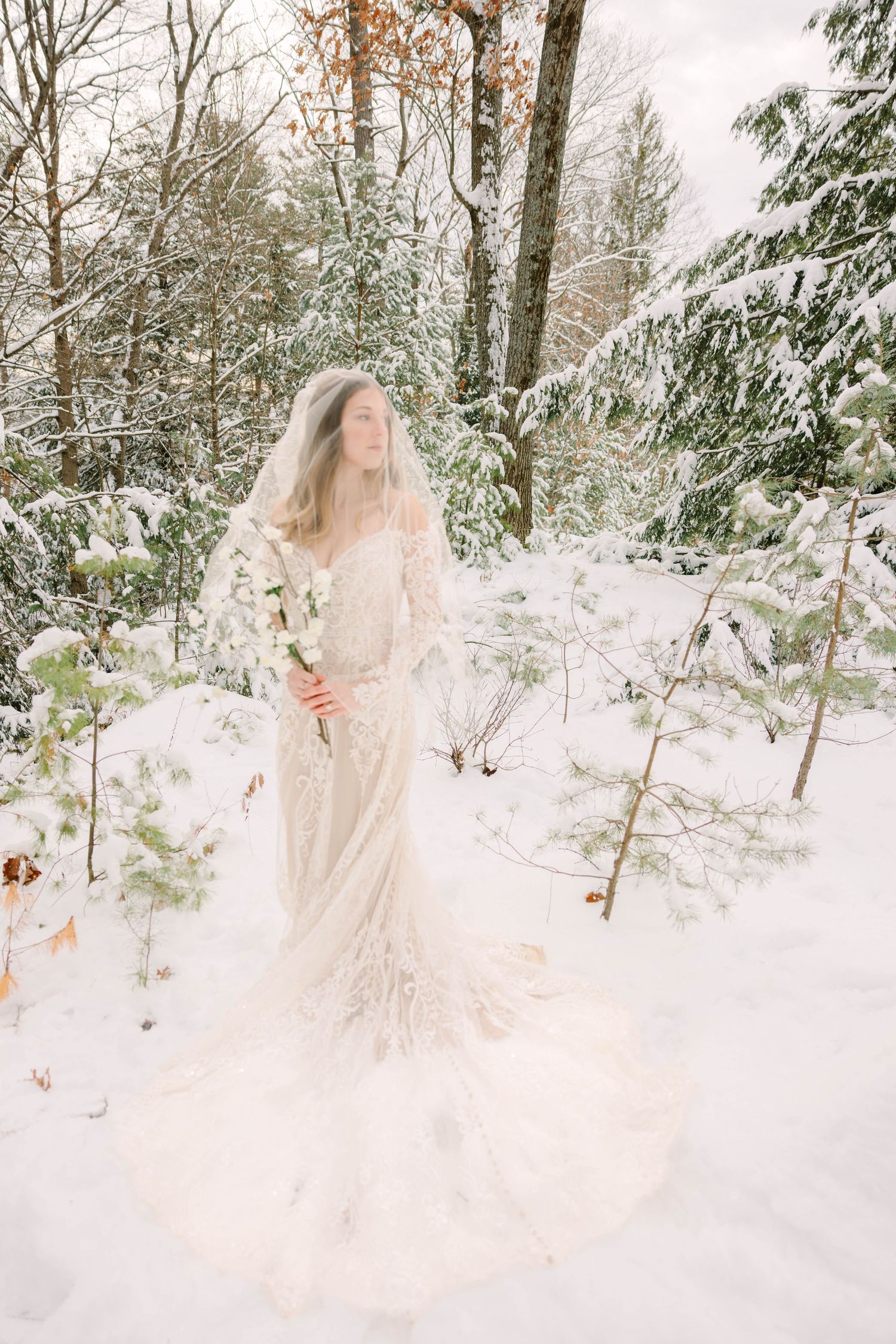 Bride in lace gown and veil holding white floral bouquet in snowy winter forest.