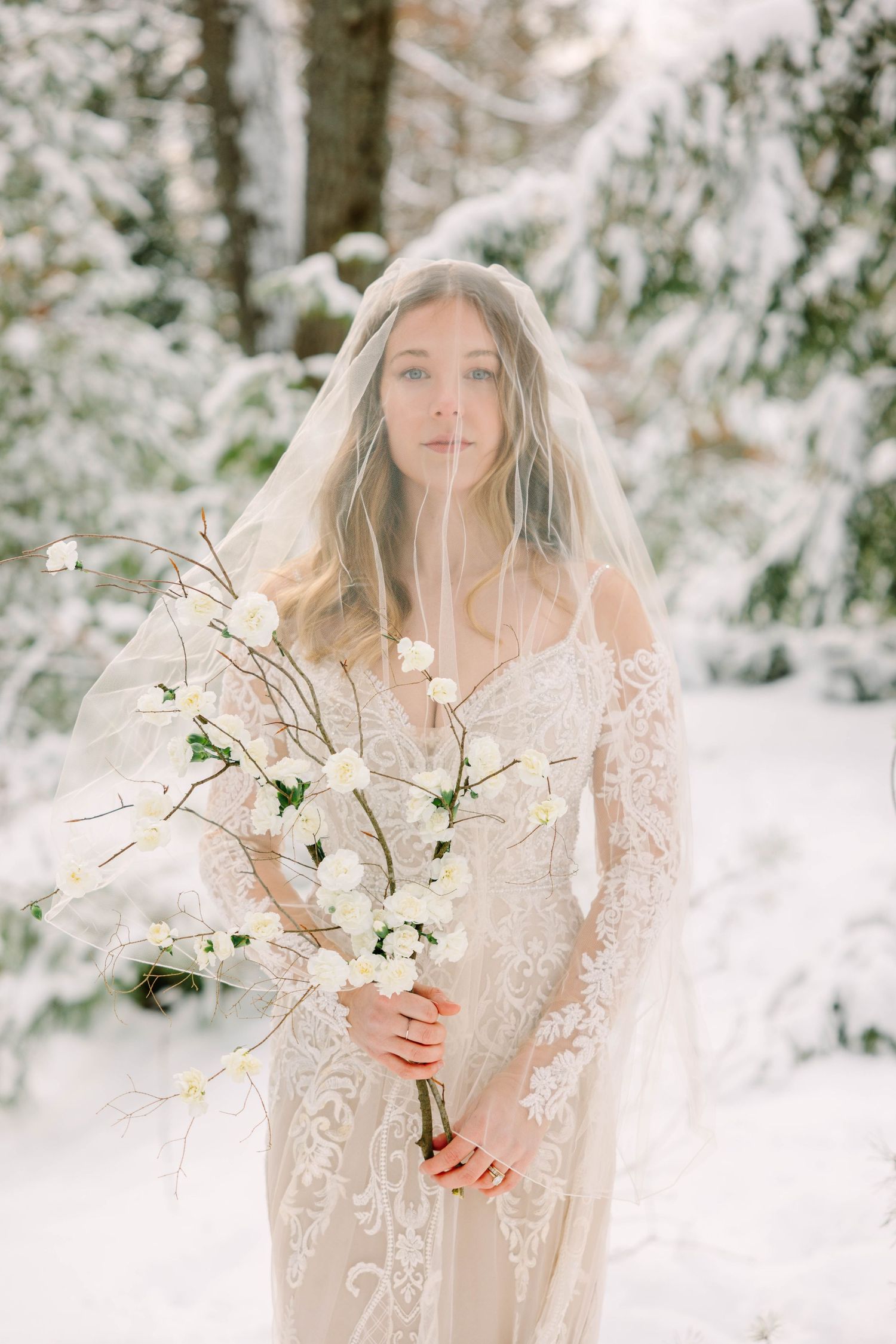 Bride in lace off-shoulder gown and veil holds branched white floral bouquet in snowy woodland setting.