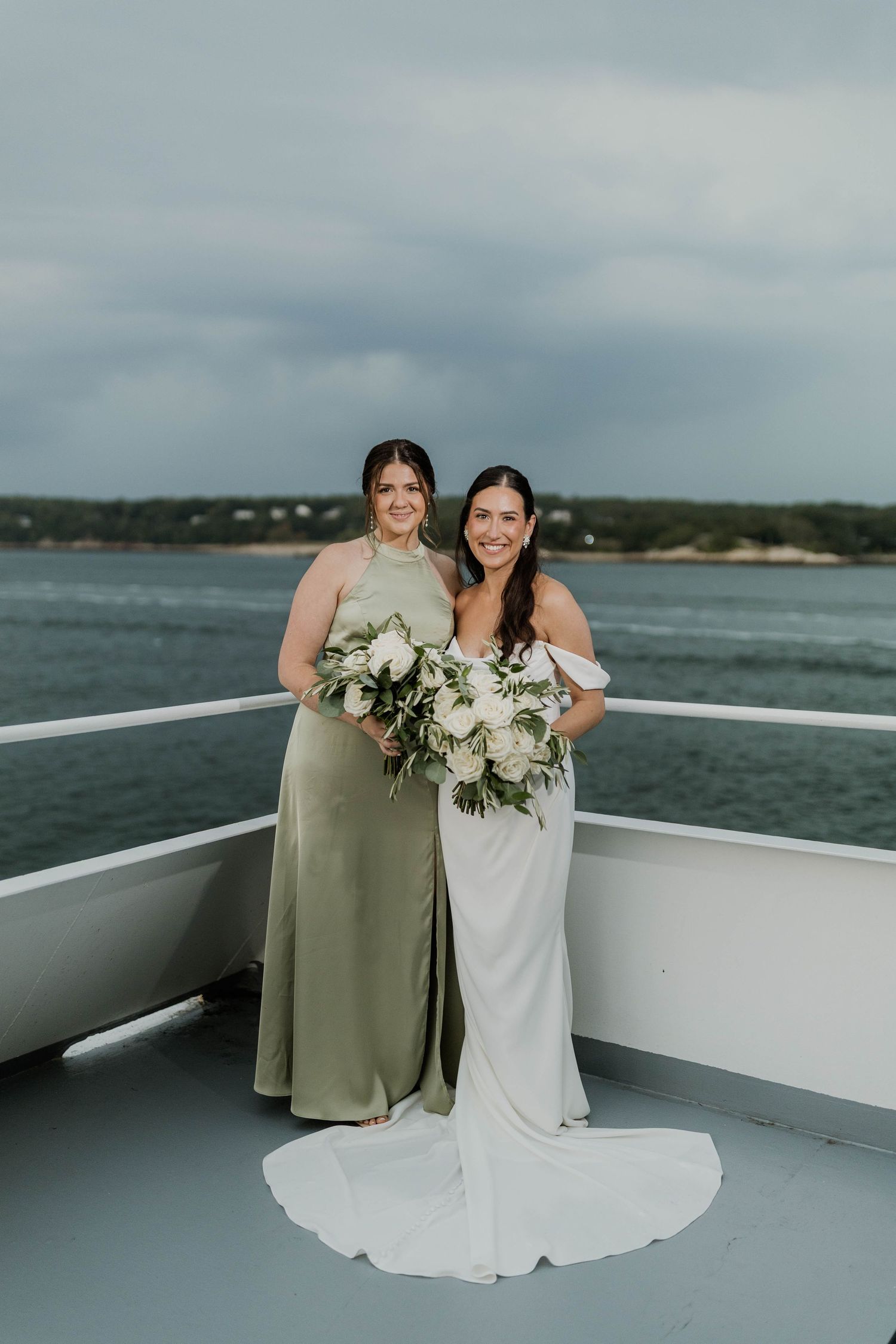 Bride in white gown poses with bridesmaid in sage green dress holding white bouquets on a boat deck.
