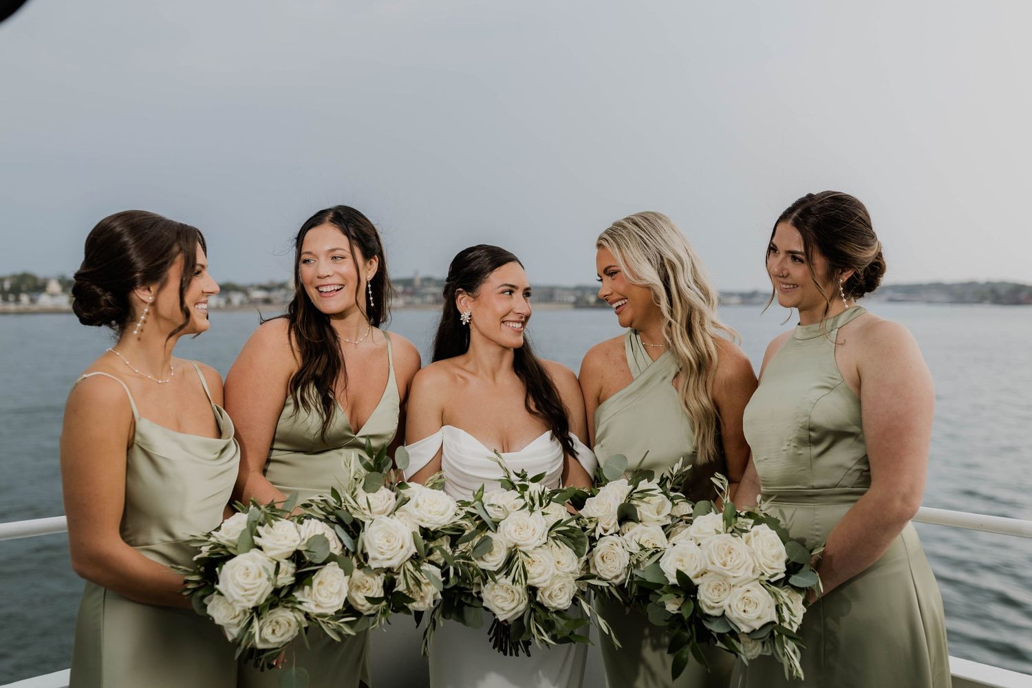 Bride with four bridesmaids in sage green dresses holding white floral bouquets on a boat on the water.
