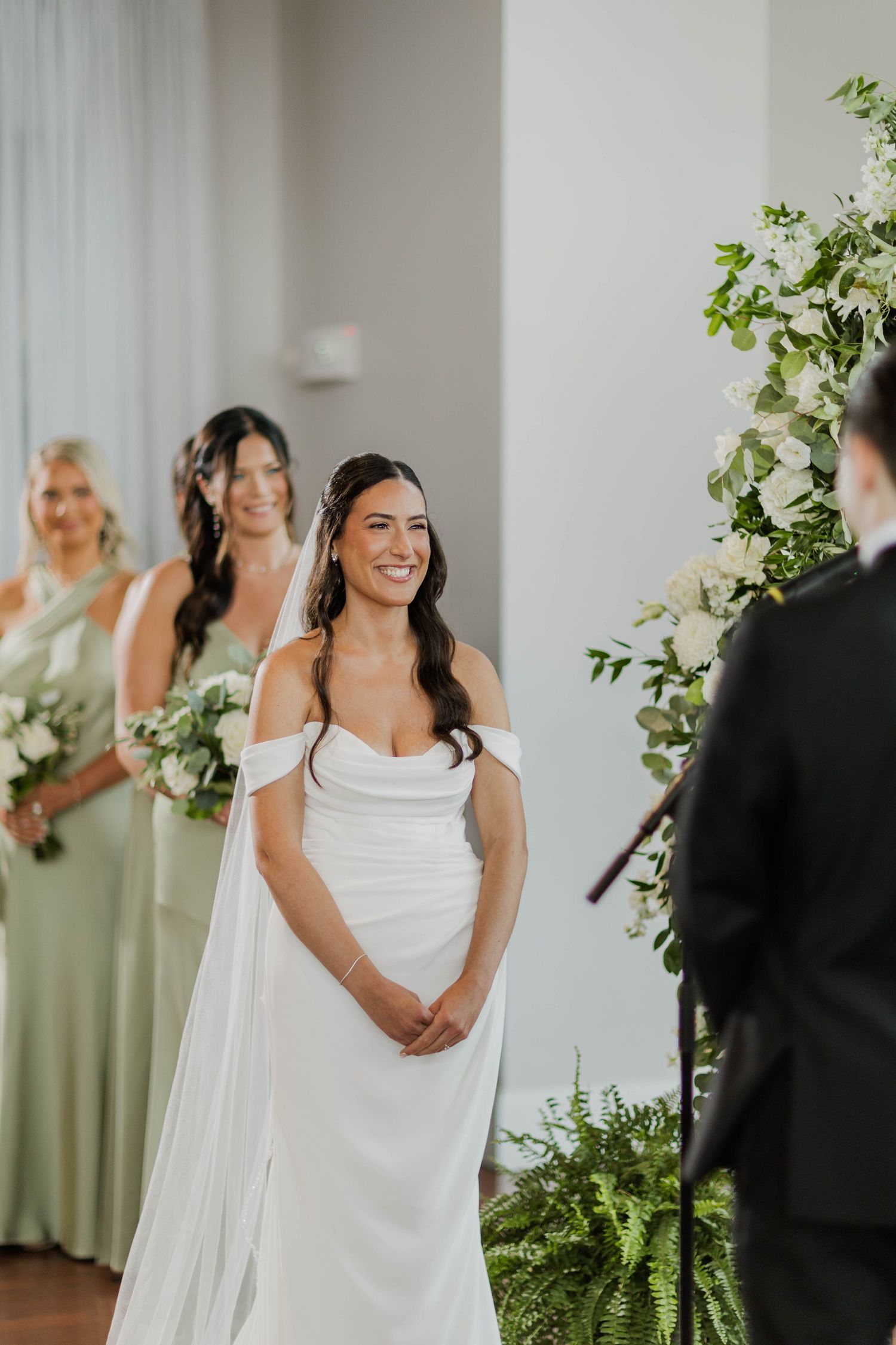 Radiant bride in white off-shoulder gown smiling at groom during wedding ceremony with bridesmaids behind.