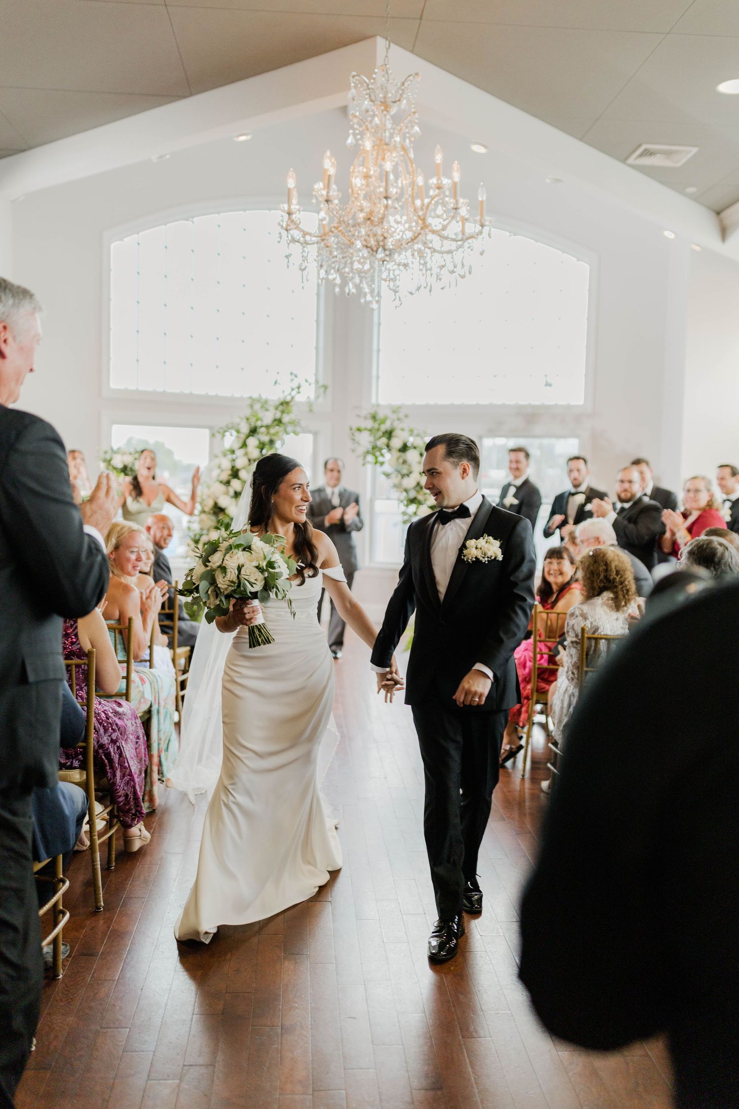 Newlyweds walking hand-in-hand down aisle after wedding ceremony under crystal chandelier.