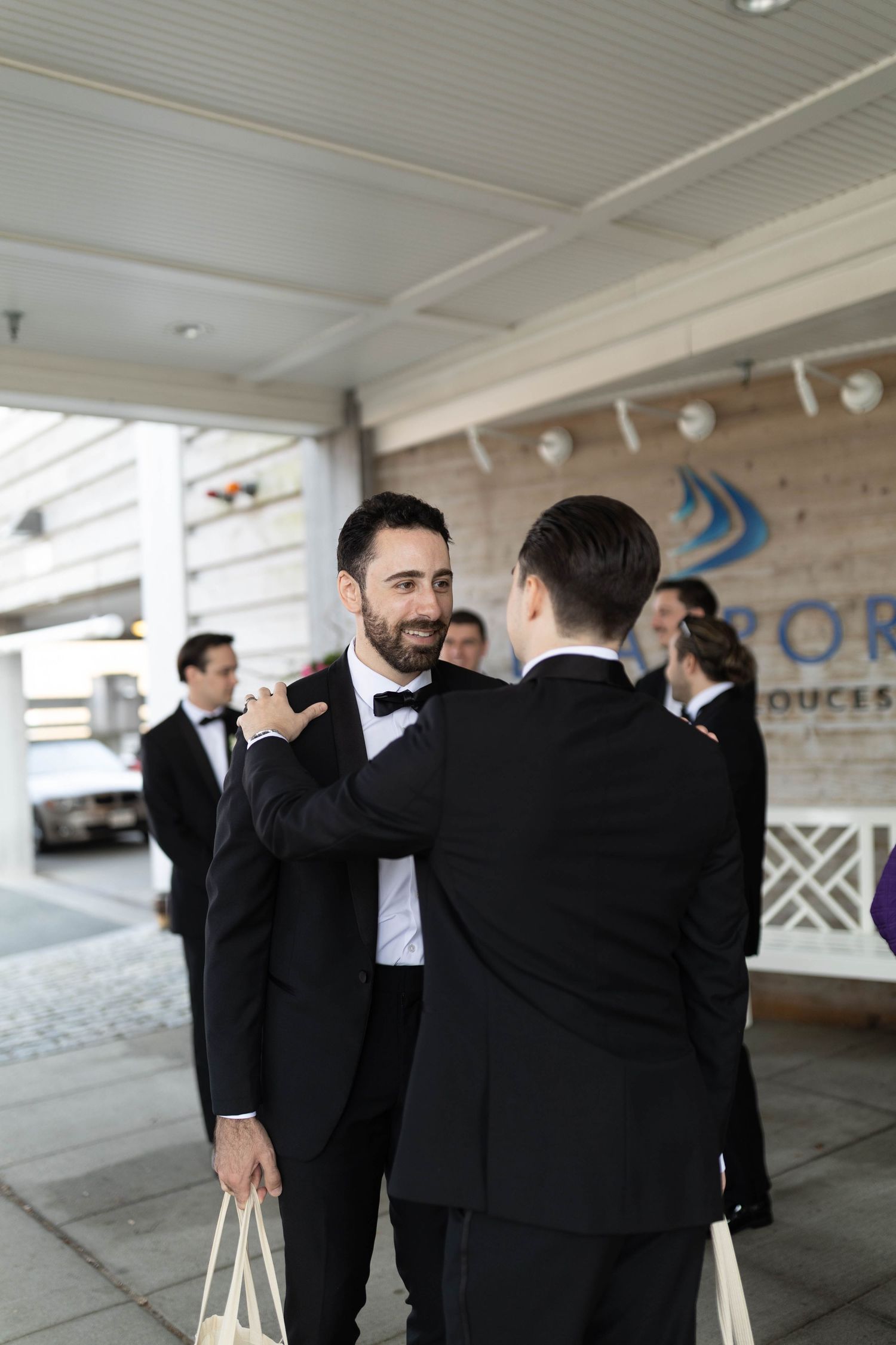 Two groomsmen in black tuxedos greeting each other outside a wedding venue.