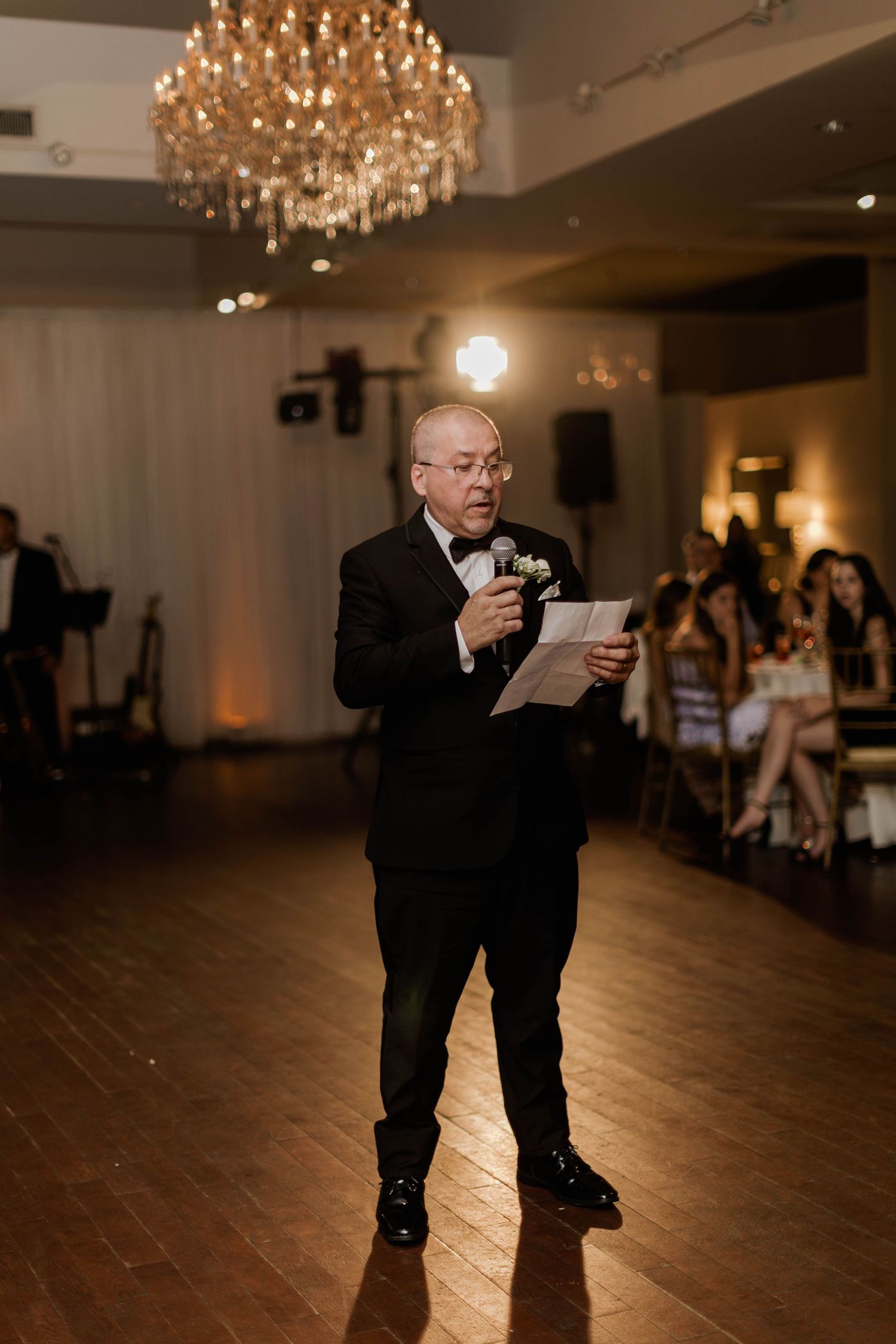 Older gentleman in tuxedo delivers heartfelt speech holding paper under crystal chandelier at wedding reception.