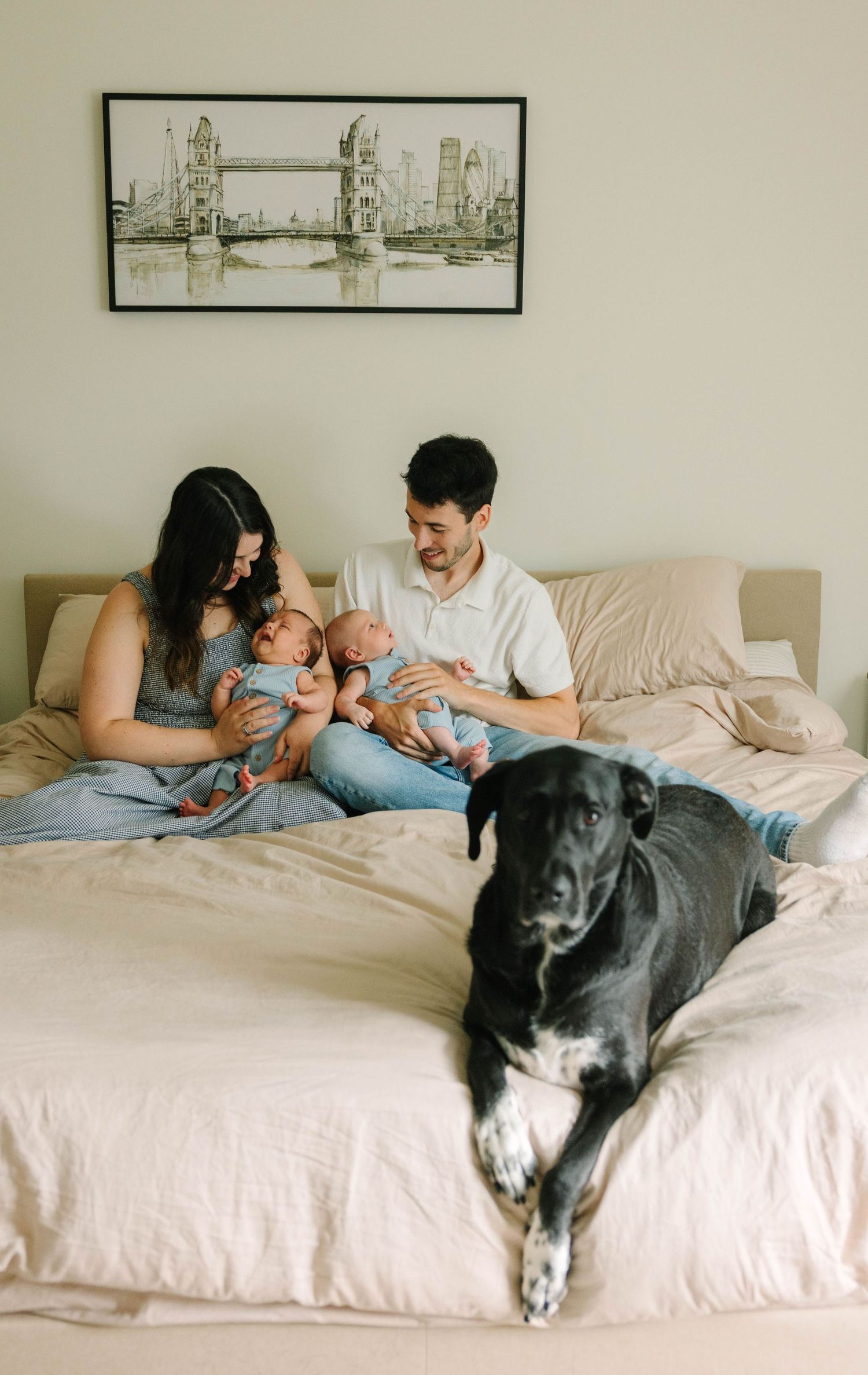 Happy parents with twin babies and black dog relaxing on bed beneath London Bridge artwork.
