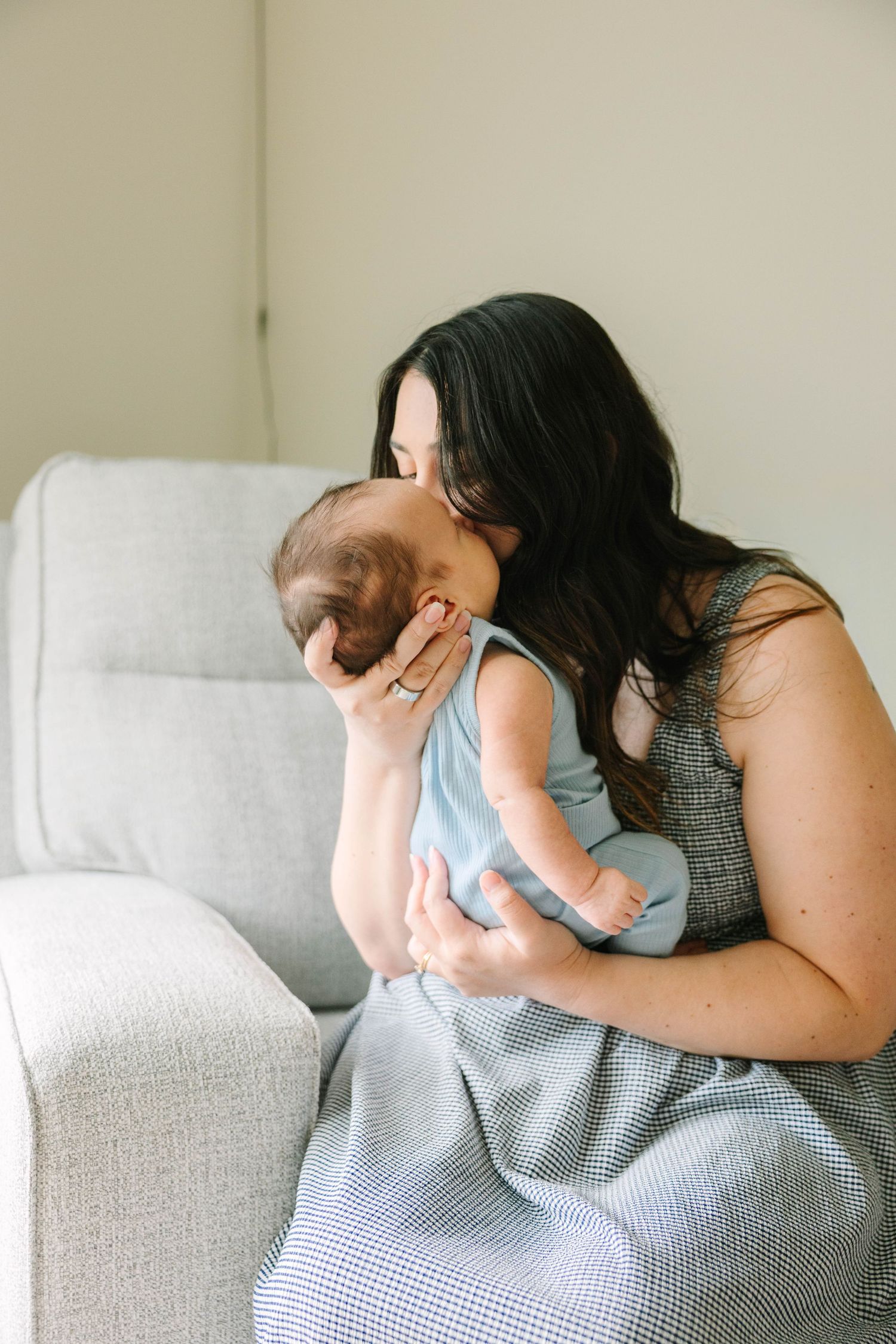 Dark-haired mother kissing her newborn baby while sitting on a gray couch in a bright room.