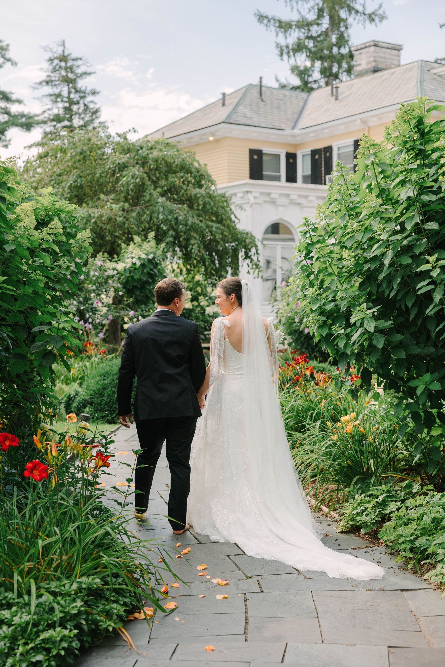 A bride and groom walk hand in hand along a stone garden path surrounded by lush greenery toward an elegant estate.