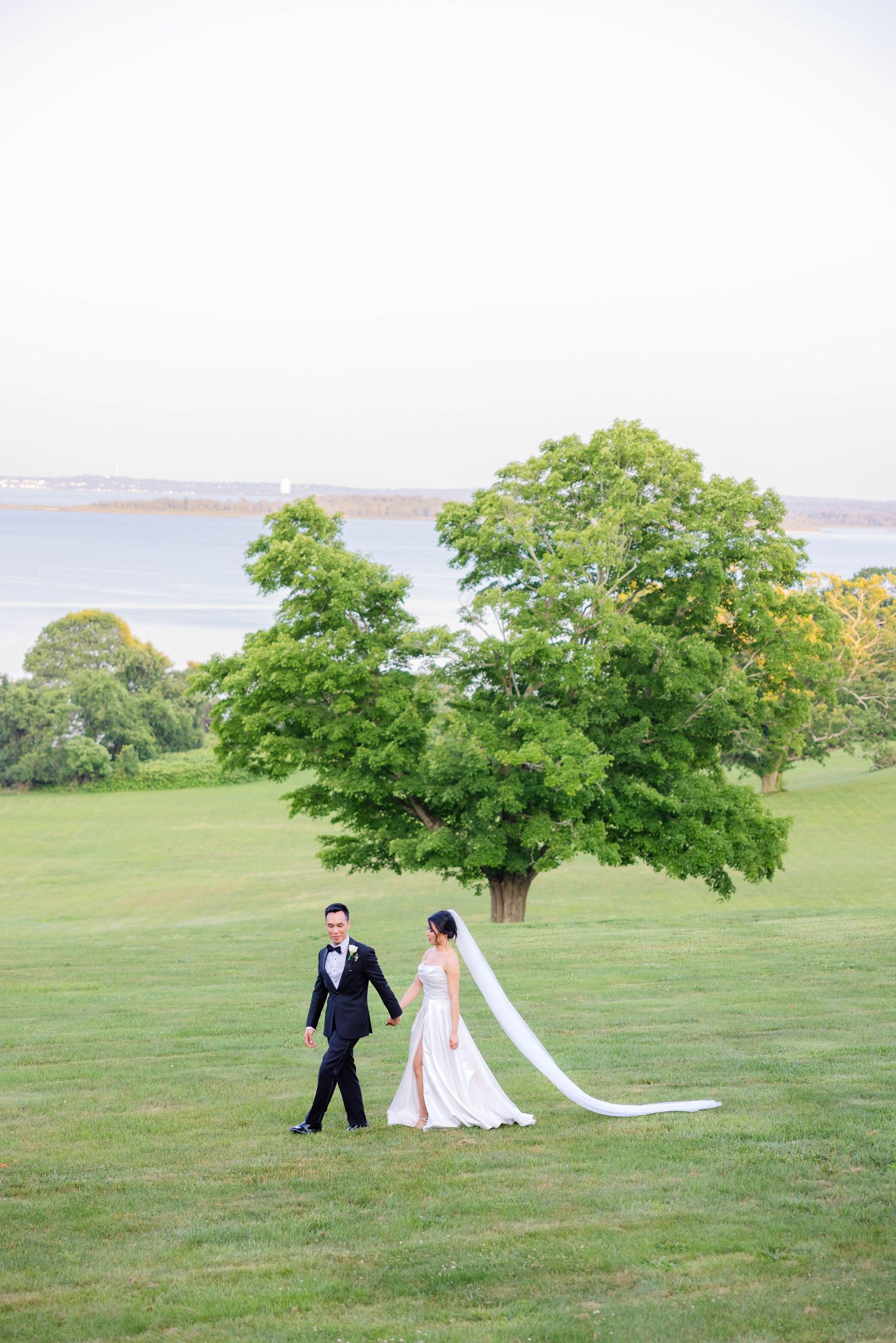 Bride and groom walk on vast green lawn near waterfront with long veil flowing in the breeze.