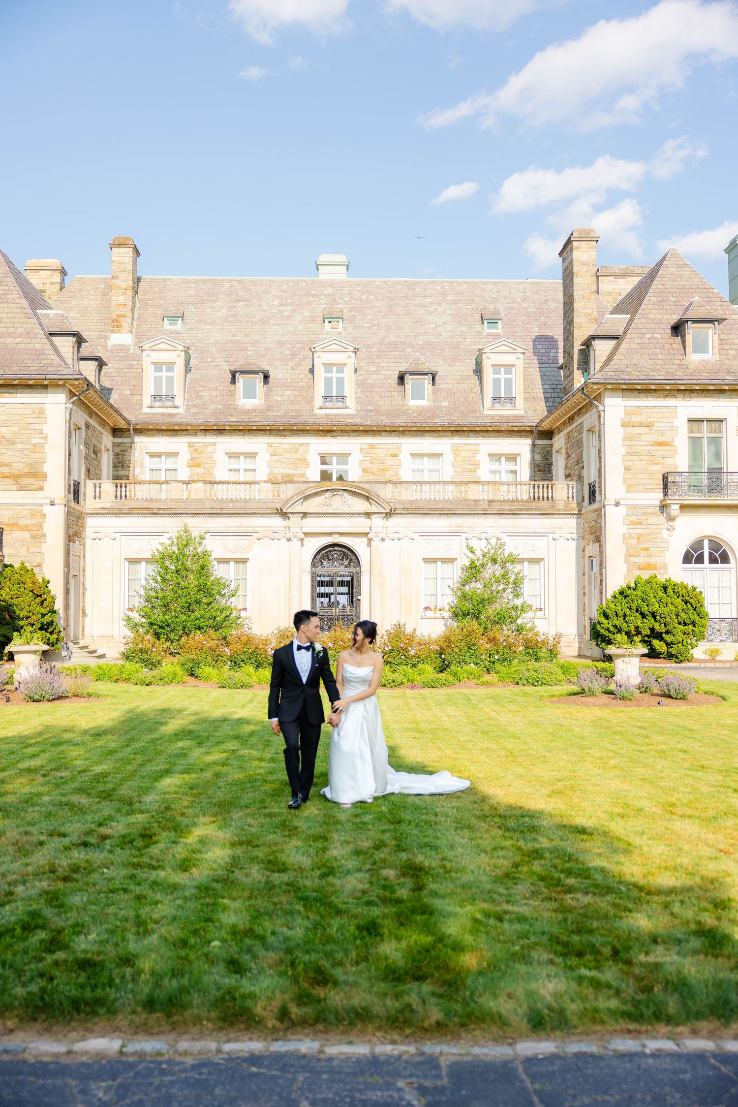 Bride and groom hold hands on green lawn in front of a grand French chateau-style estate.