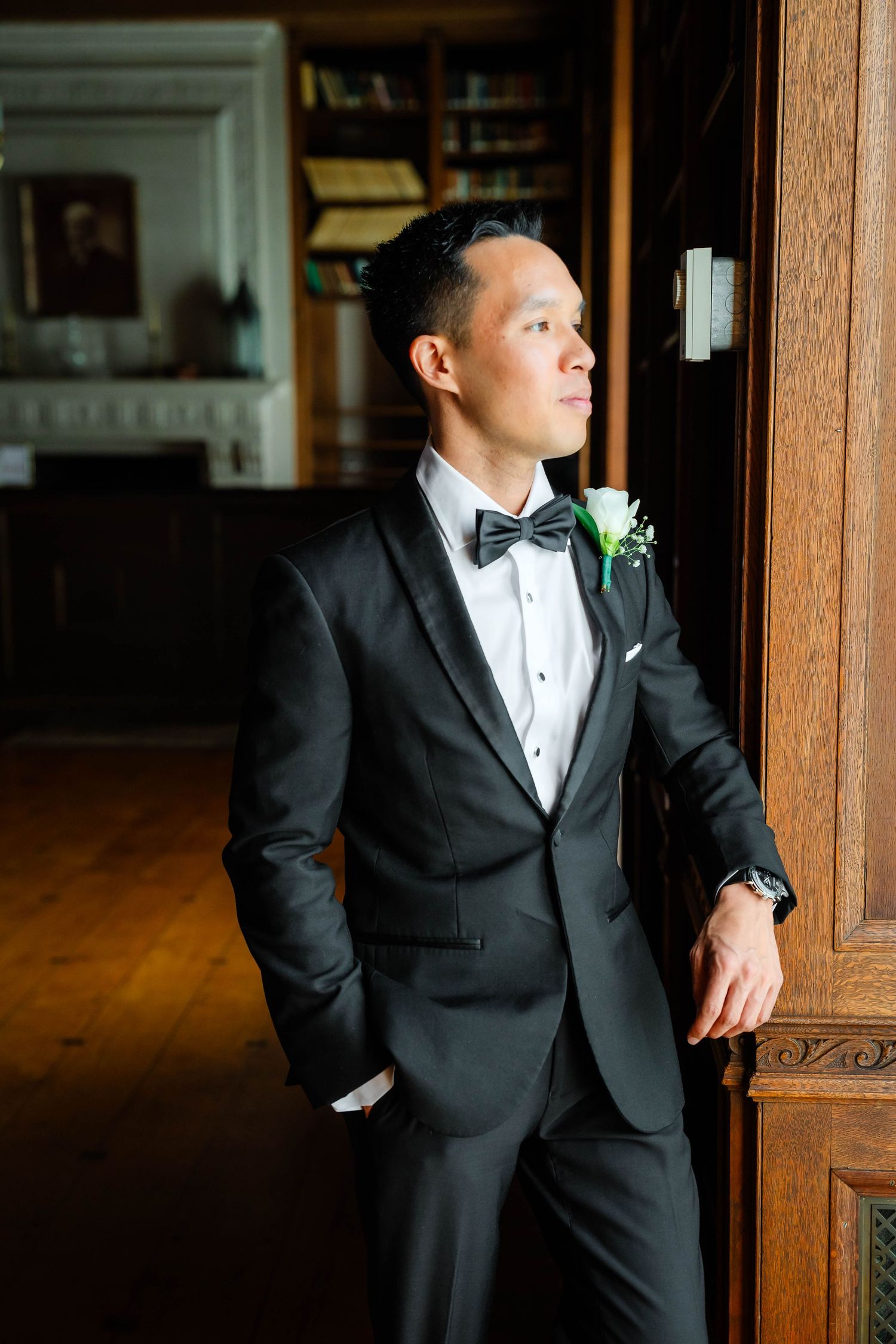 Dapper groom in black tuxedo with bow tie and boutonniere leaning against a wooden door.