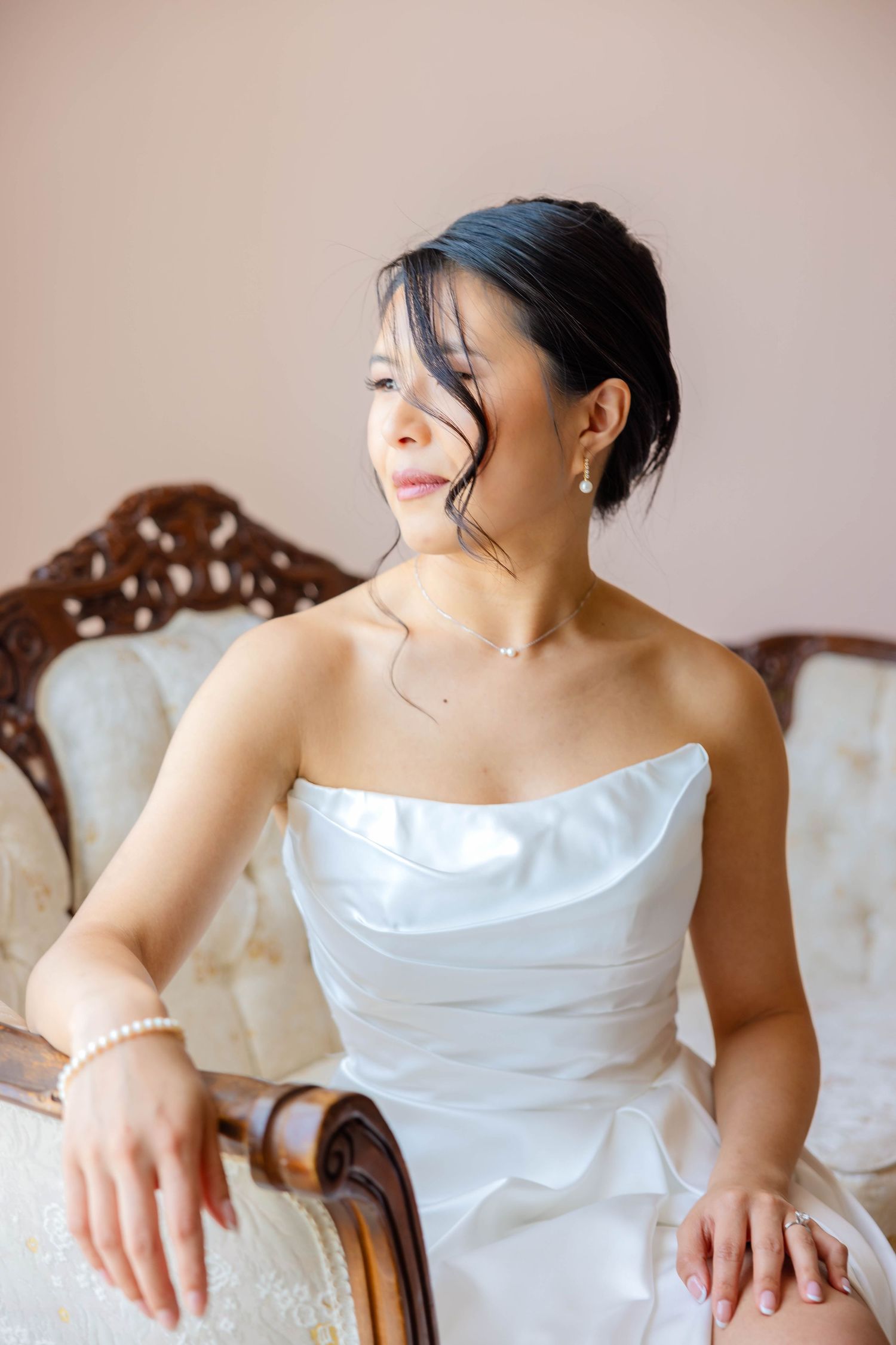 Elegant bride in strapless white gown sits on vintage chair wearing pearl jewelry.