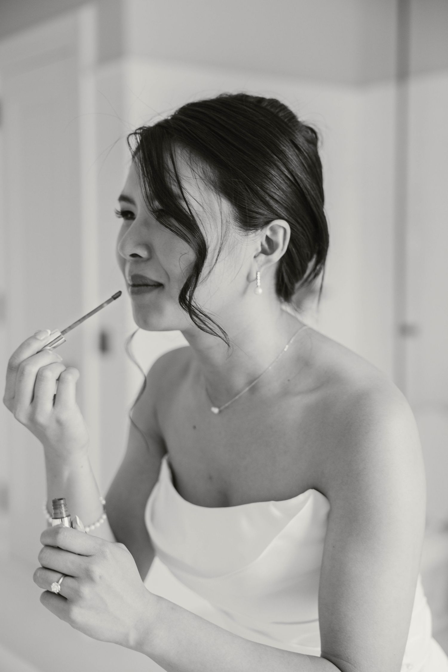 Bride in white strapless dress applying lipstick while getting ready in black and white photo.