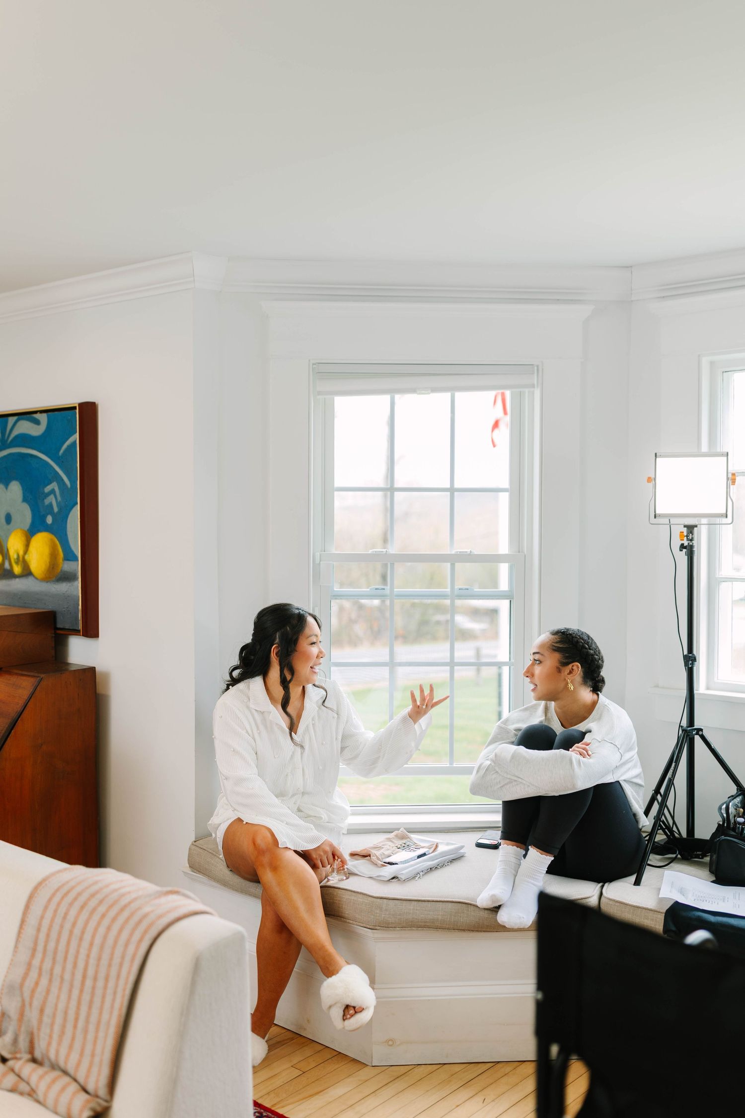 Bride and friend in robes chatting on a window seat during wedding morning preparations.