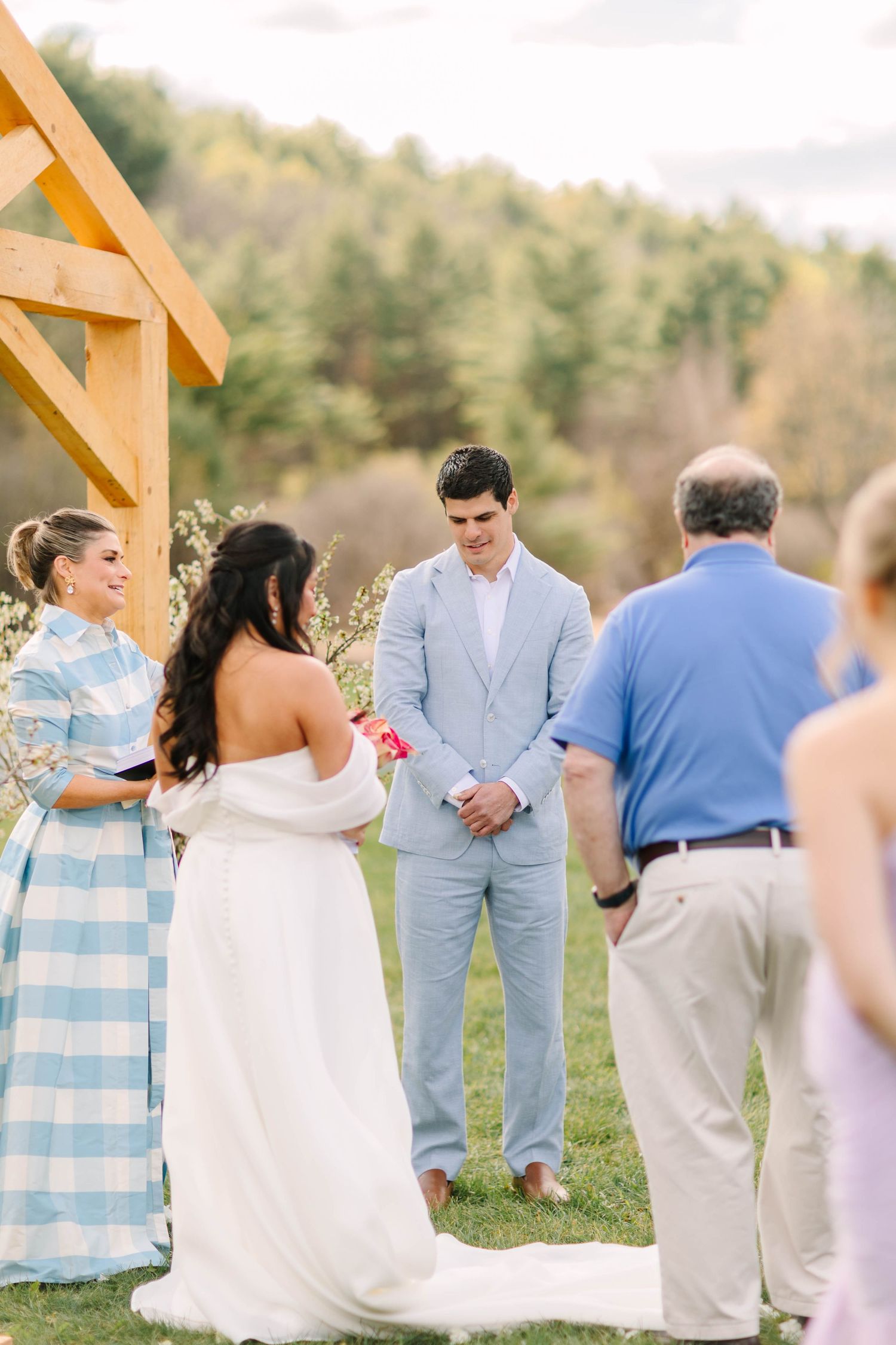 Outdoor wedding ceremony with bride in white gown and groom in light blue suit exchanging vows near wooden arbor.