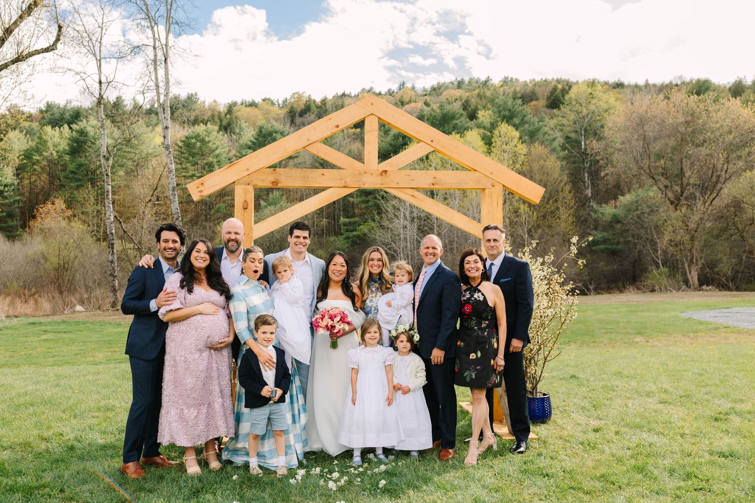 Smiling wedding family group photo under rustic wooden arch with fall foliage hillside in background.
