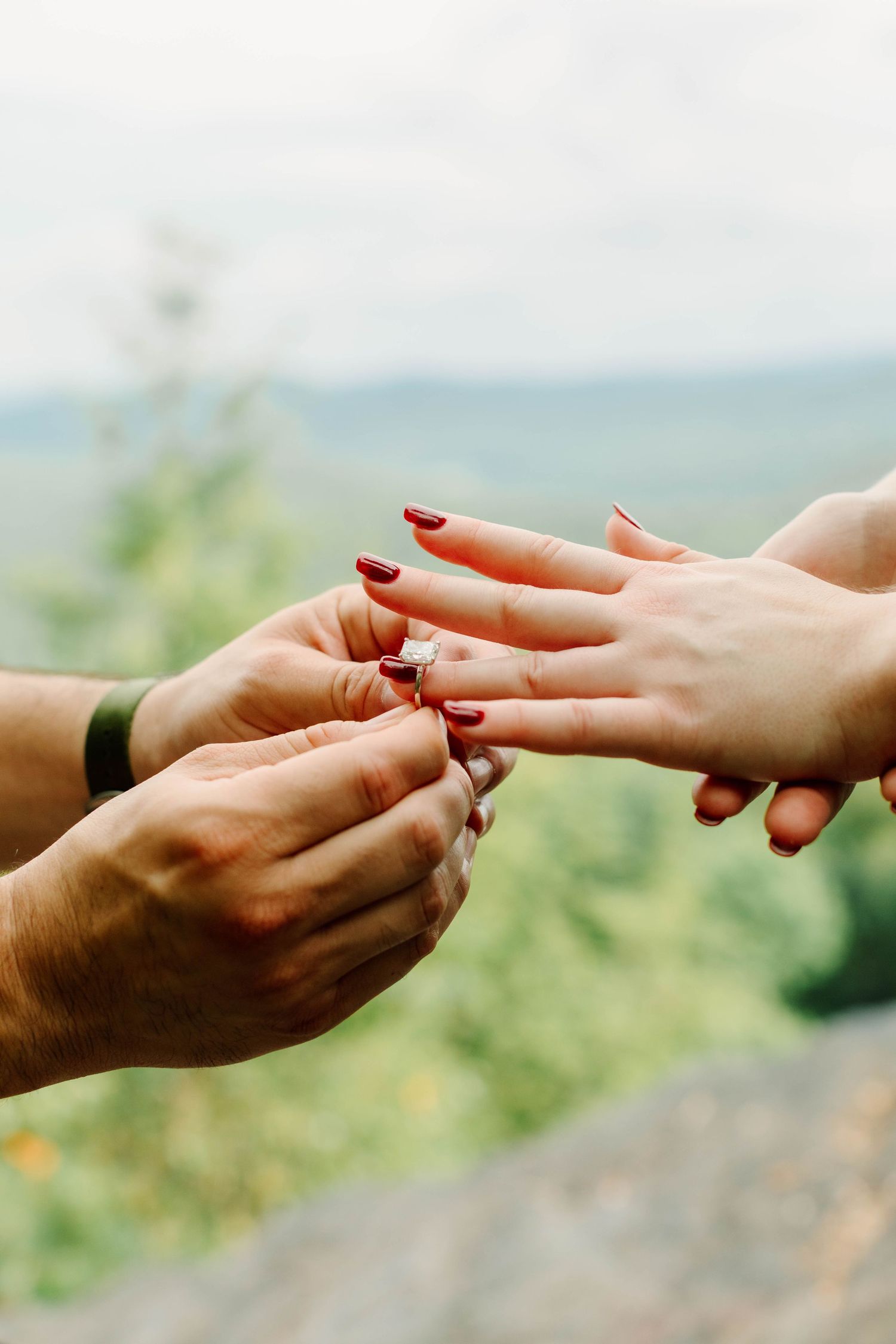 A man placing a diamond engagement ring on a woman's finger with red nails, outdoors with a scenic green backdrop.