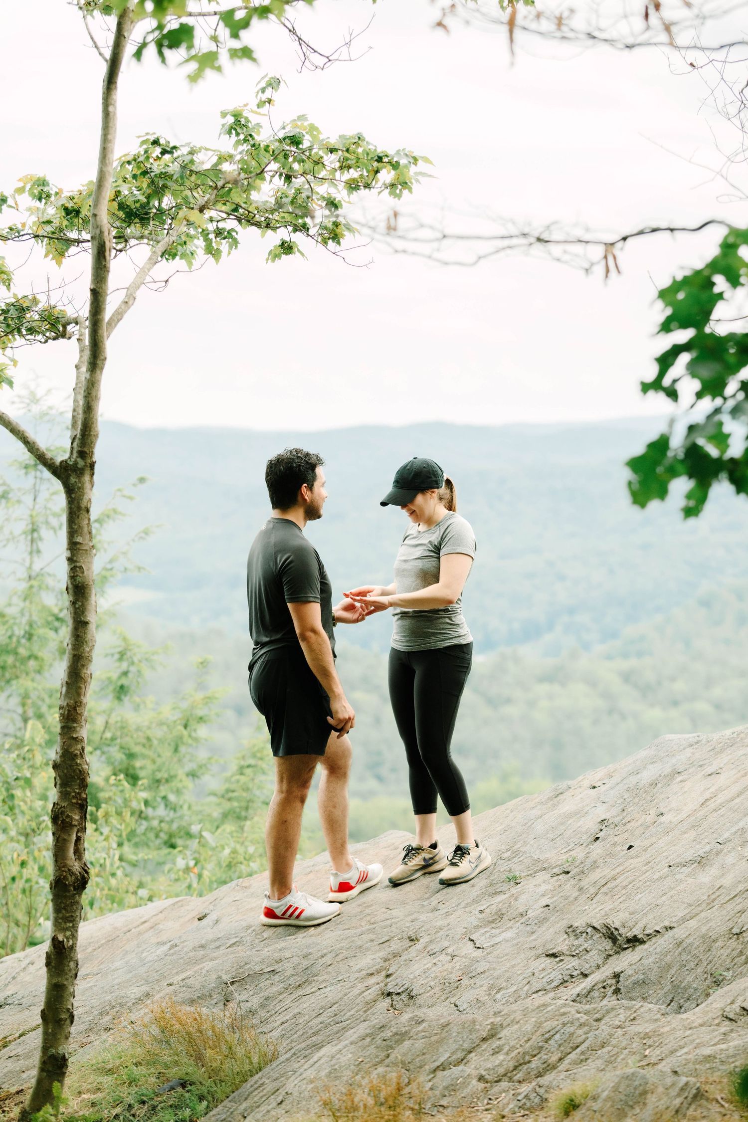 Couple examining an engagement ring on a rocky mountain overlook surrounded by trees.