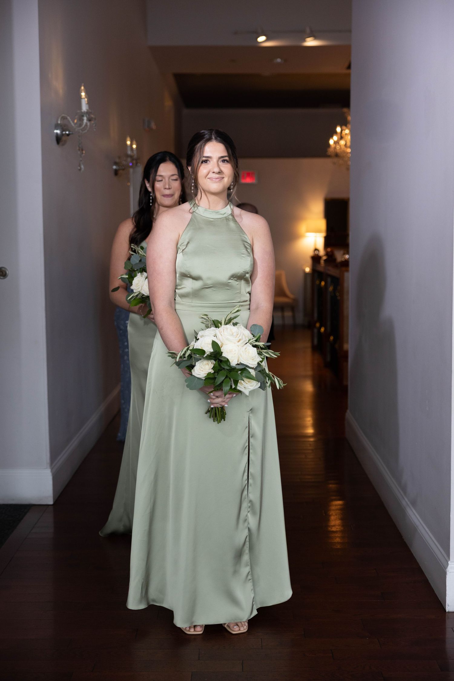 Two bridesmaids in sage green satin gowns carrying white bouquets walking down a hallway.