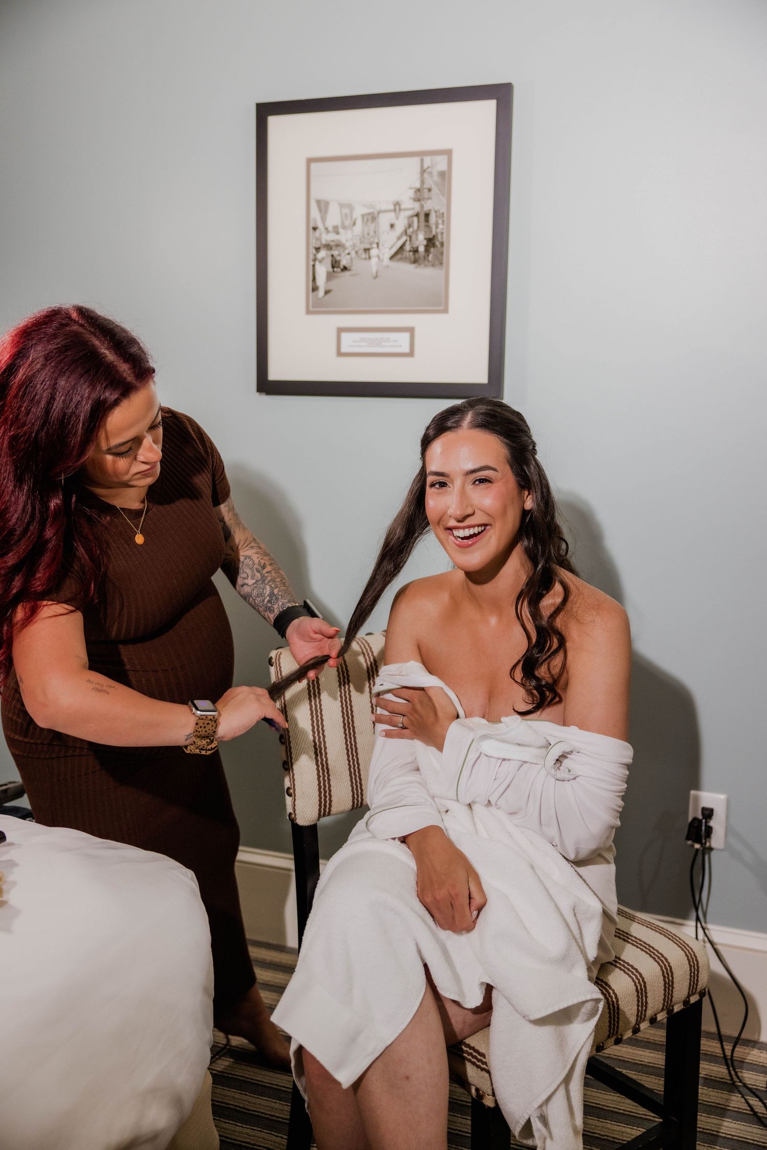 Bride in white robe laughing while getting hair done by stylist in hotel room before wedding.