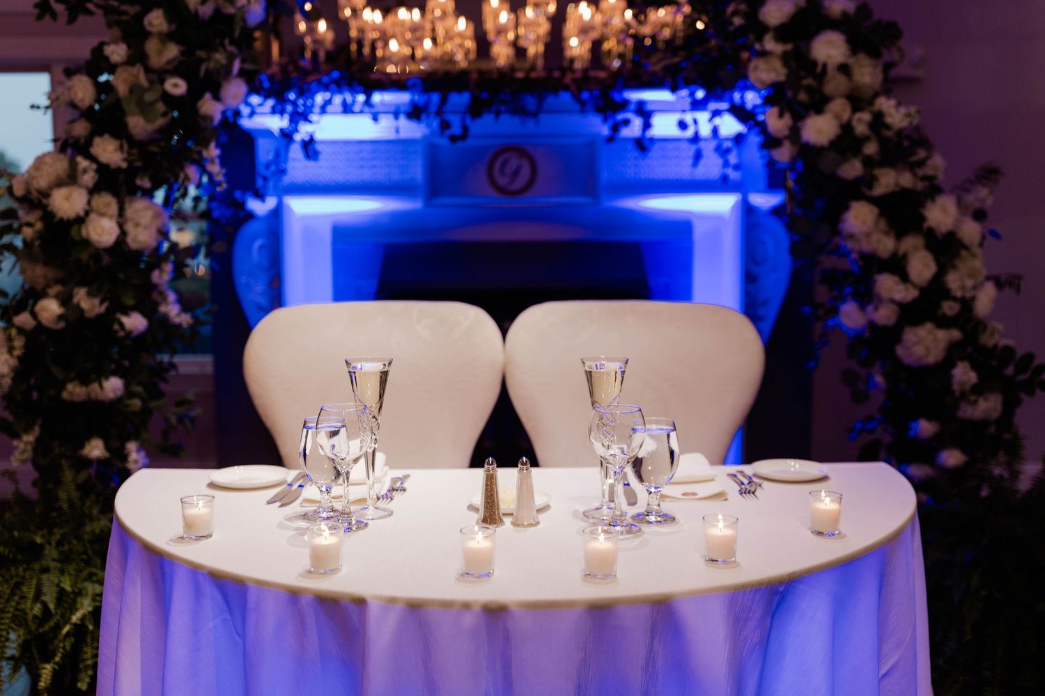 Elegant wedding sweetheart table with white chairs, candlelight, and floral arch with blue uplighting.