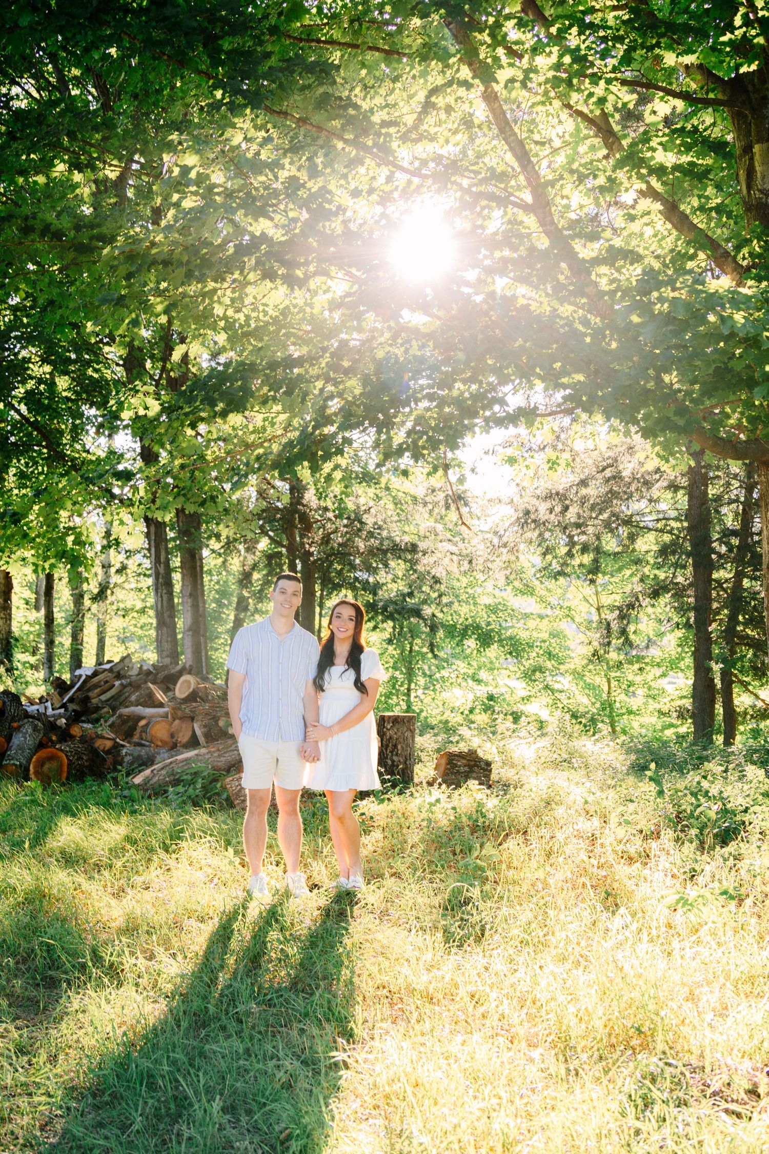 Couple in white outfits standing in sunlit forest clearing during golden hour.