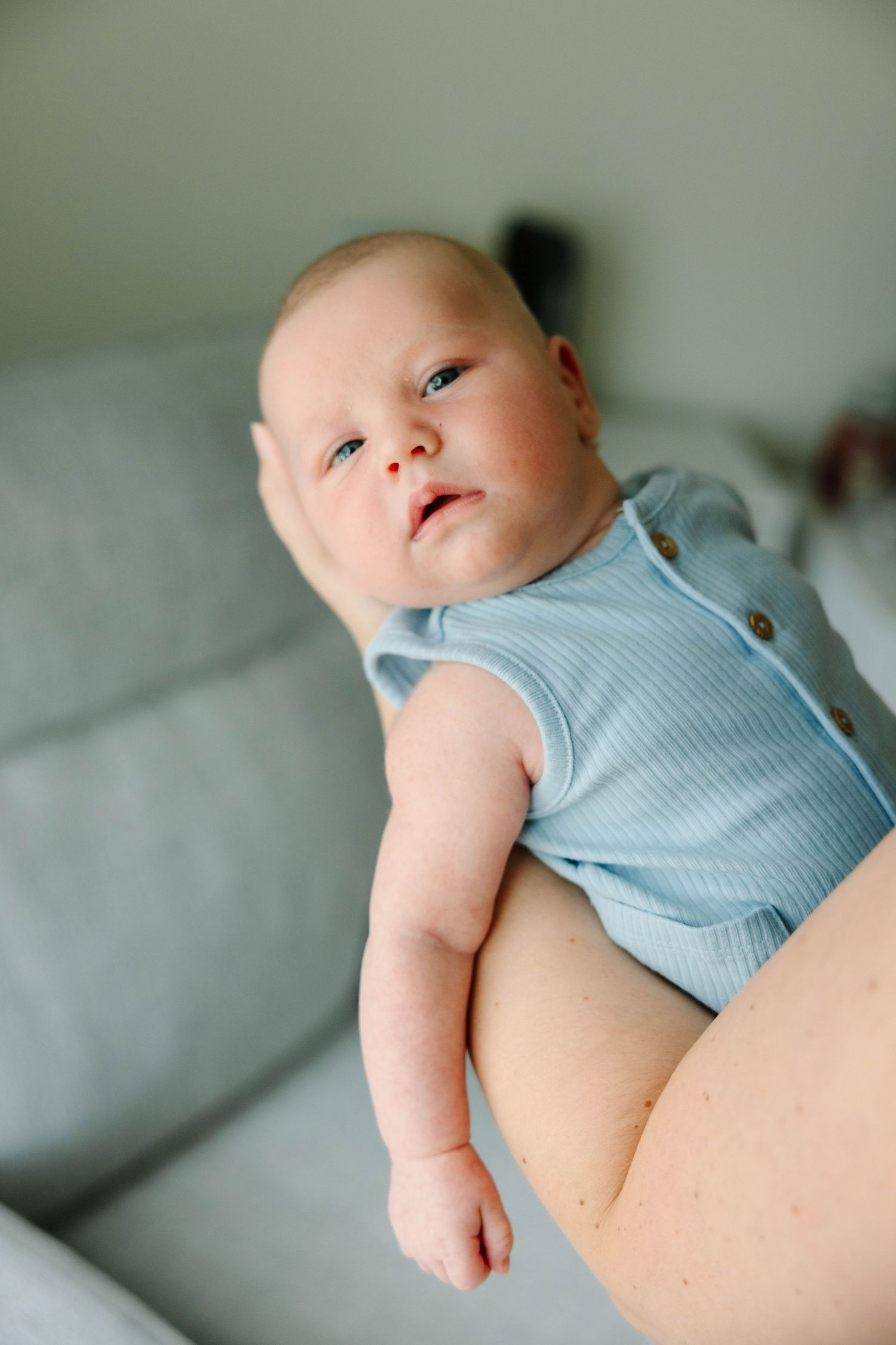 Adorable newborn baby in light blue ribbed onesie being held by mother on a gray sofa.