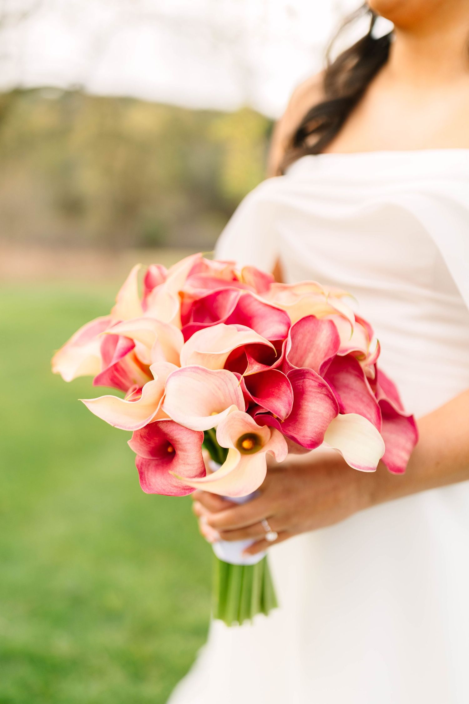 Close-up of bride holding elegant pink and cream calla lily wedding bouquet in white off-shoulder dress.