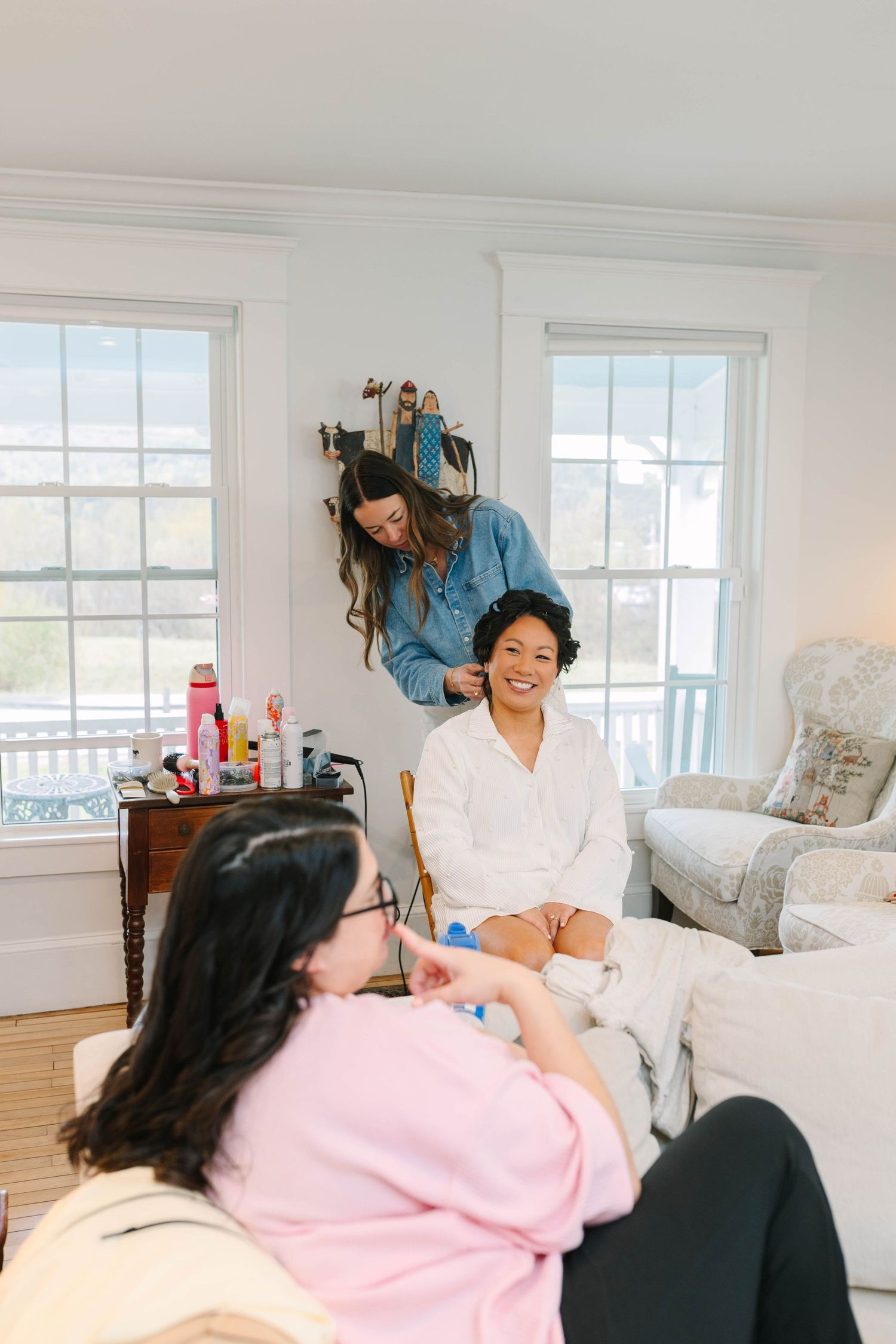 Bride in white robe smiles while hairstylist works during wedding morning preparations.