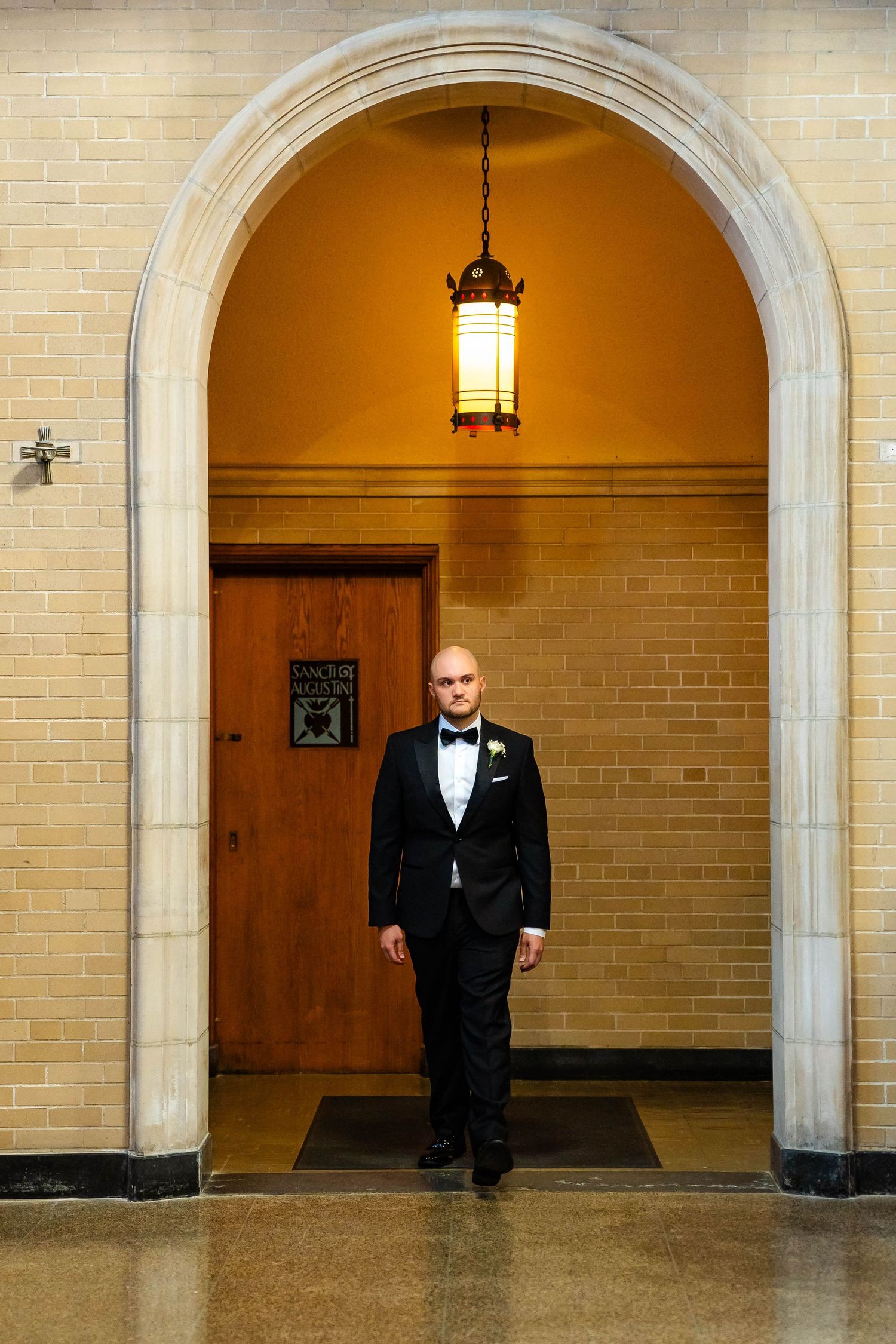 Groom in black tuxedo stands alone under glowing lantern in stone church archway.