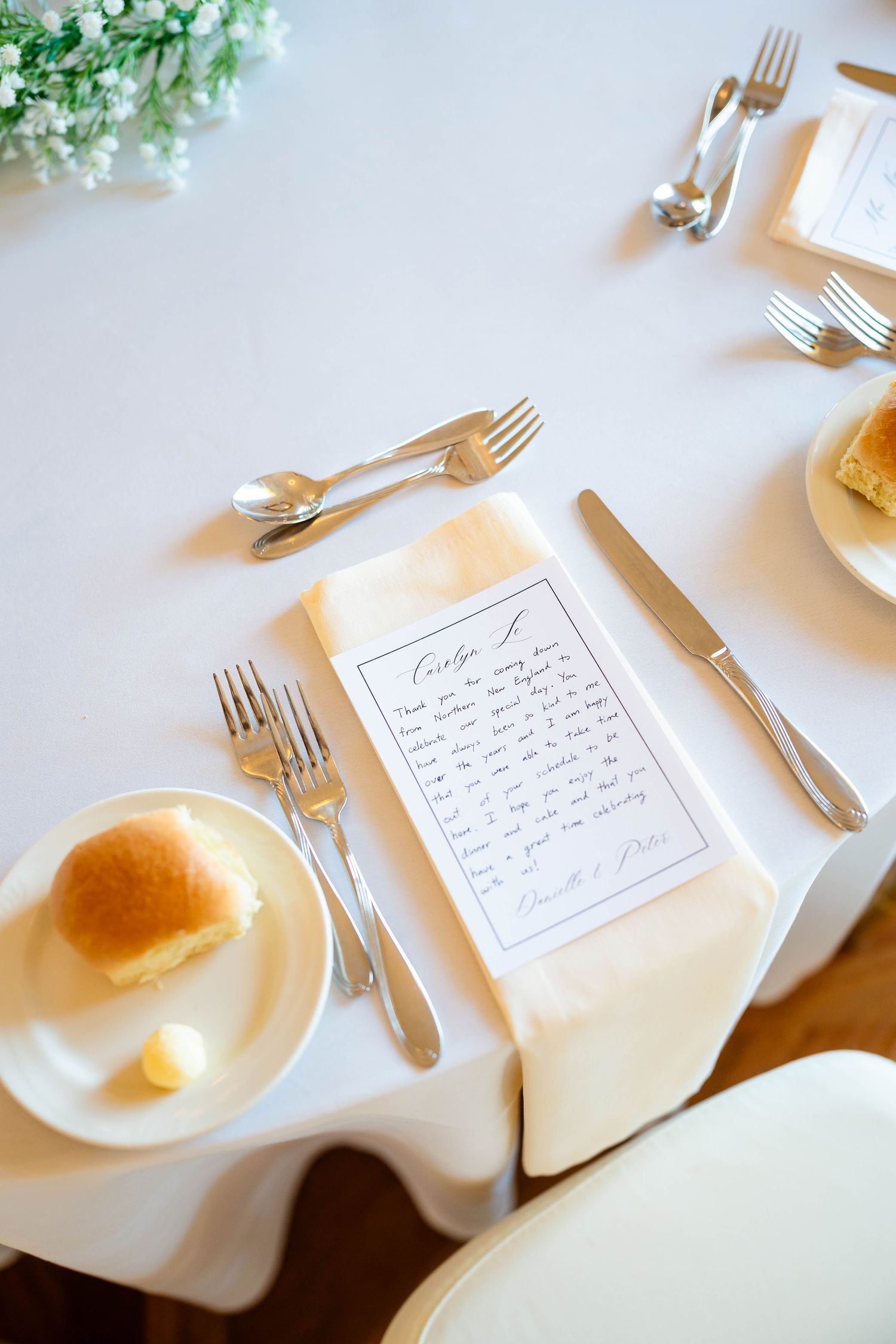 Elegant wedding place setting with handwritten menu card, silverware, bread roll, and white linens.