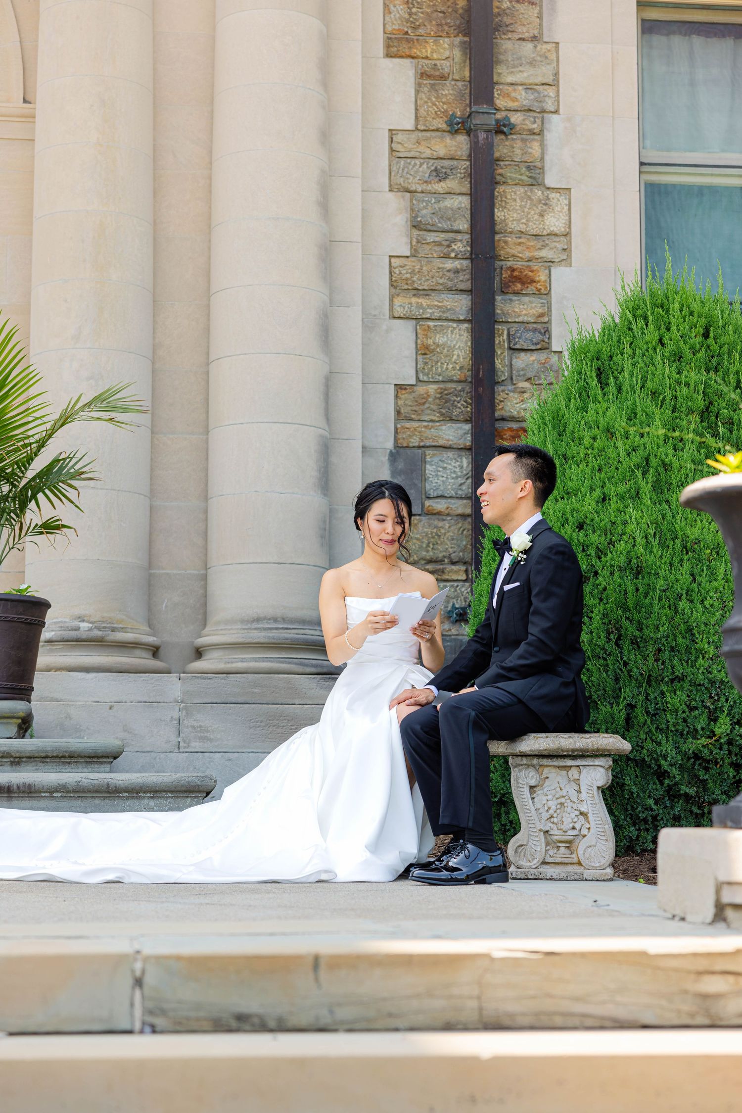 Asian bride and groom share private vow reading moment seated on stone bench outside grand columned building.