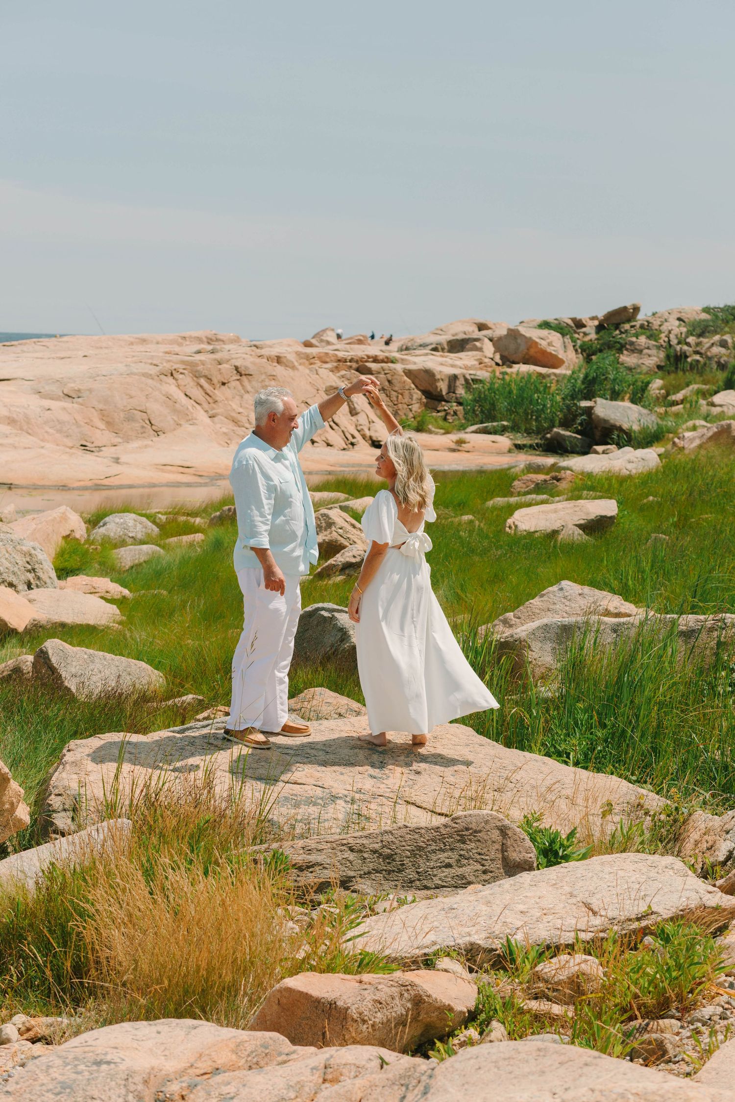 Couple in white attire standing on rocky coastal landscape with green grass and ocean views.