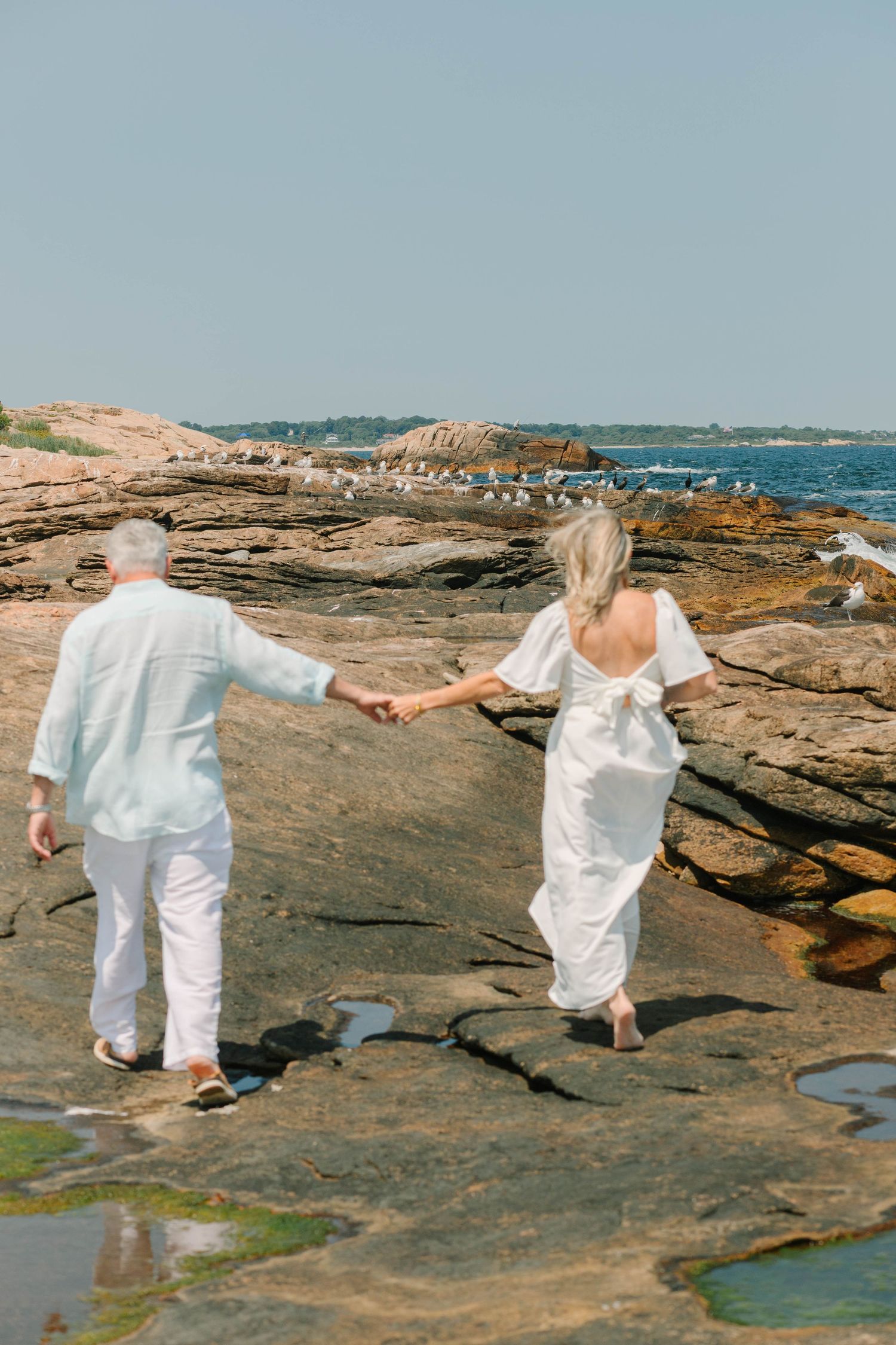 Couple in white attire holding hands while walking on rocky coastal shoreline with seagulls and ocean in background.