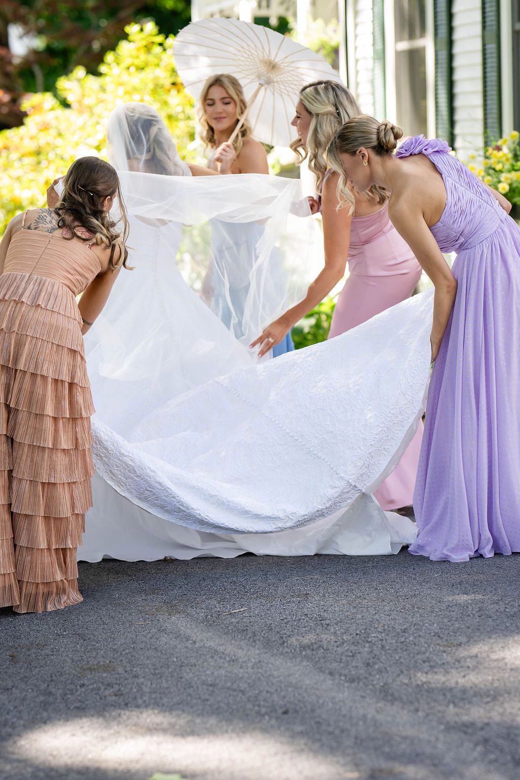 Bridesmaids in pink, lavender, and tan dresses fluff bride's white lace gown and veil outdoors.