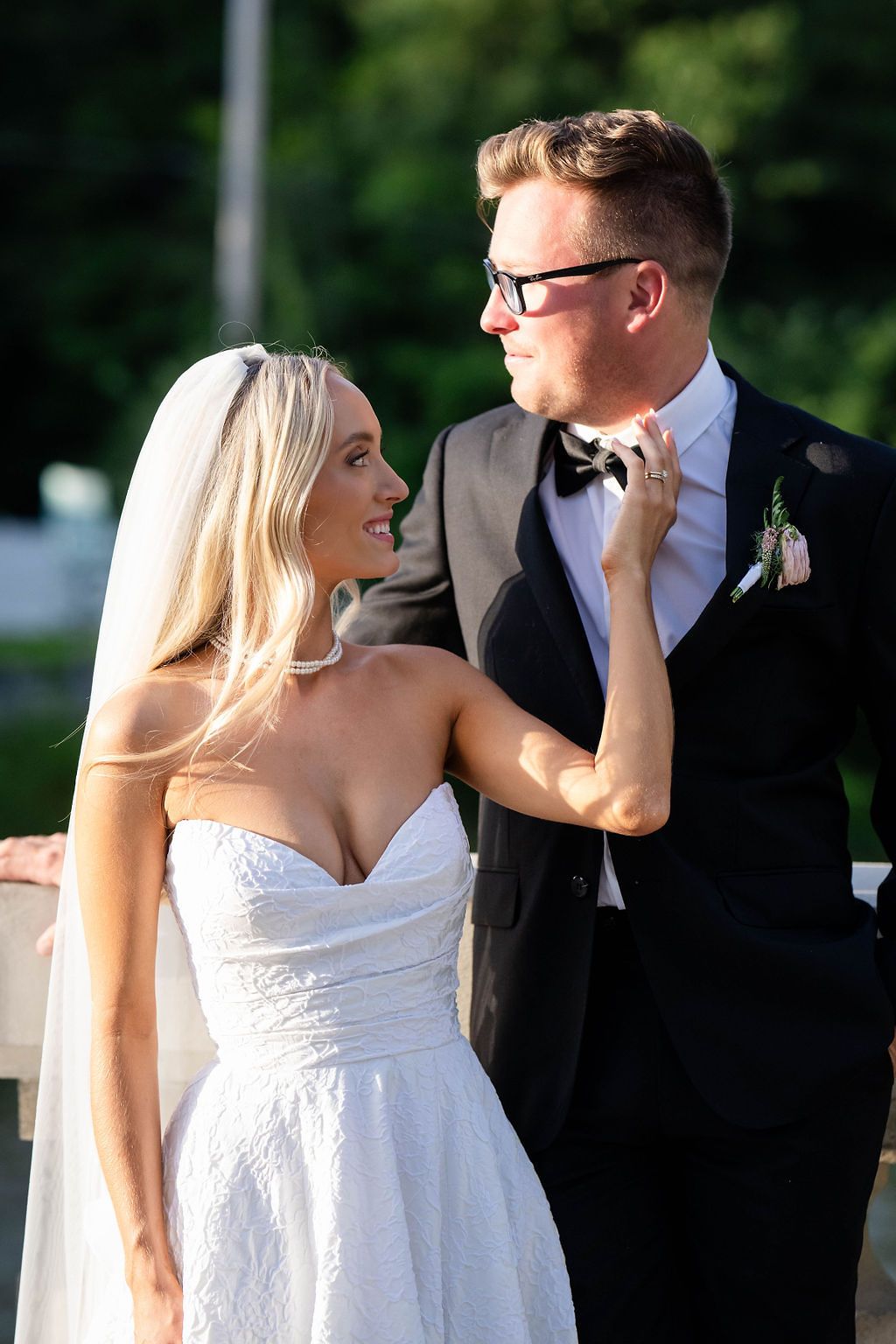 Smiling bride in strapless lace gown adjusts grooms bow tie outdoors on wedding day.