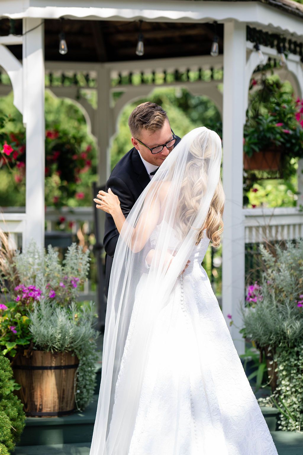Groom embracing bride in lace gown with veil near floral gazebo during first look.