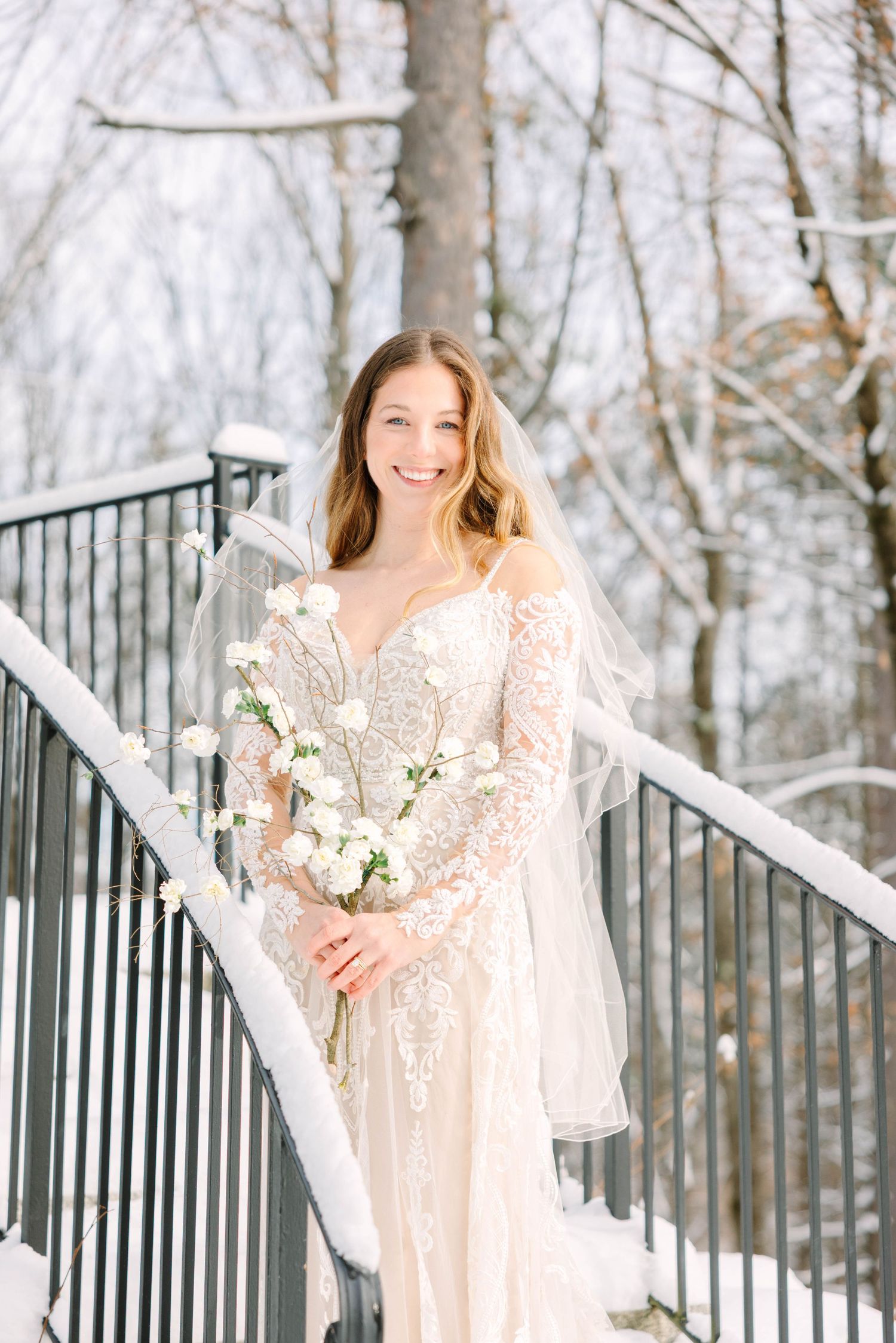 Smiling bride in lace wedding dress with veil on snow-covered staircase.