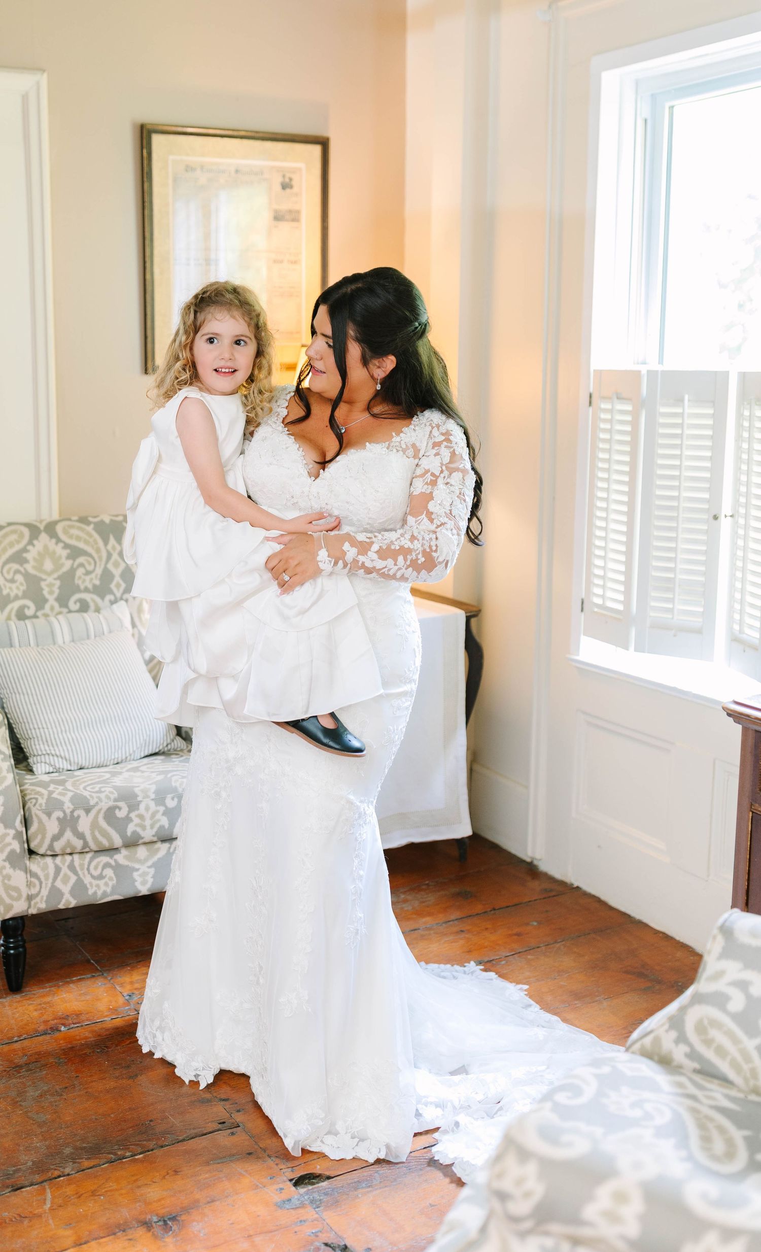 Bride in lace gown holding young flower girl by sunny window.
